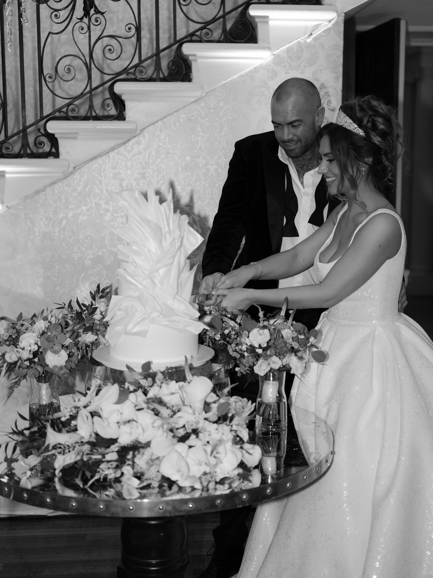 Black and white photo of a couple cutting their wedding cake decorated with flowers beside an ornate spiral staircase.