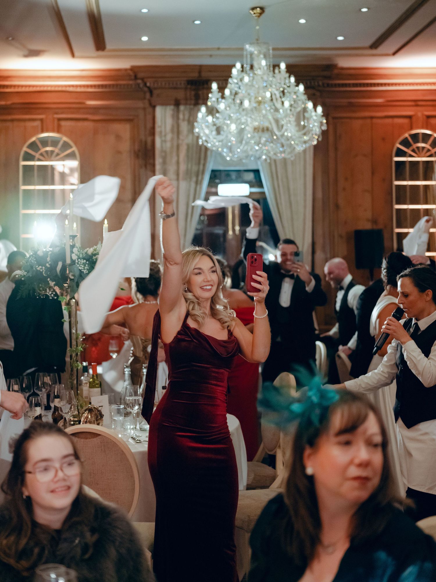 Party guests celebrate at an elegant reception with raised glasses under a crystal chandelier in a wood-paneled ballroom.