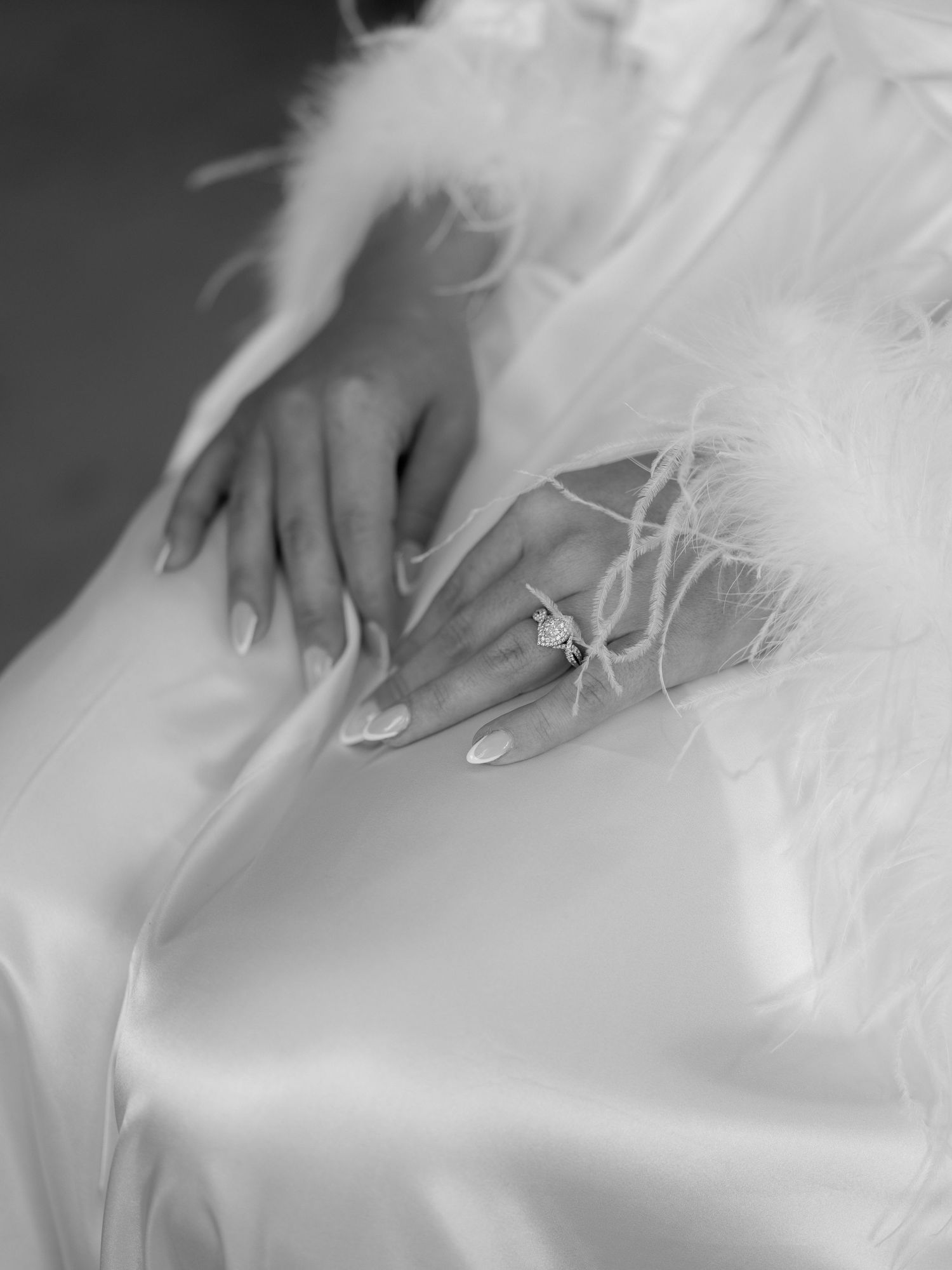 A delicate hand with manicured nails rests on white tulle fabric, wearing an elegant engagement ring.