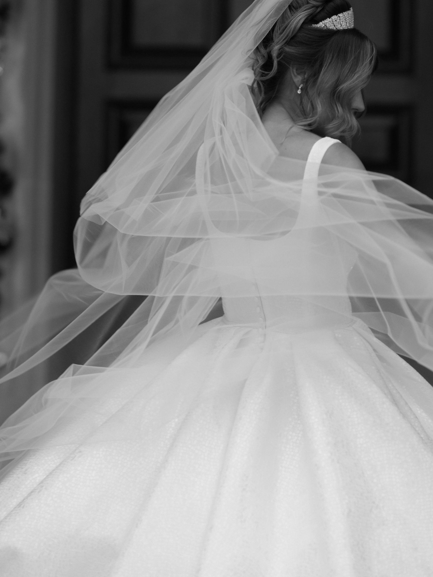 A bride wearing an elegant flowing white wedding gown with a delicate tulle veil captured in black and white photography.