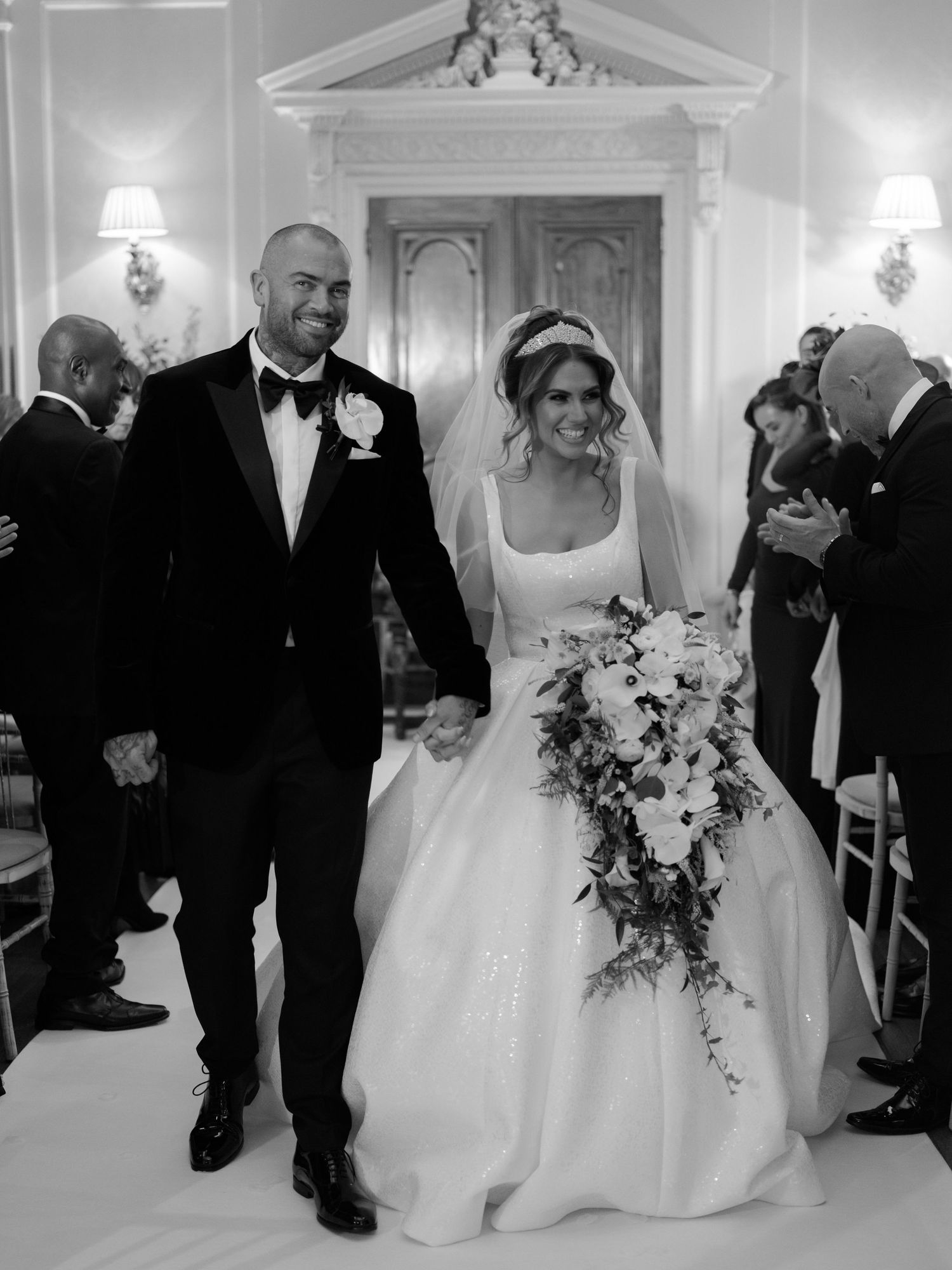 Black and white wedding photo showing a newlywed couple walking down the aisle with a cascading floral bouquet.