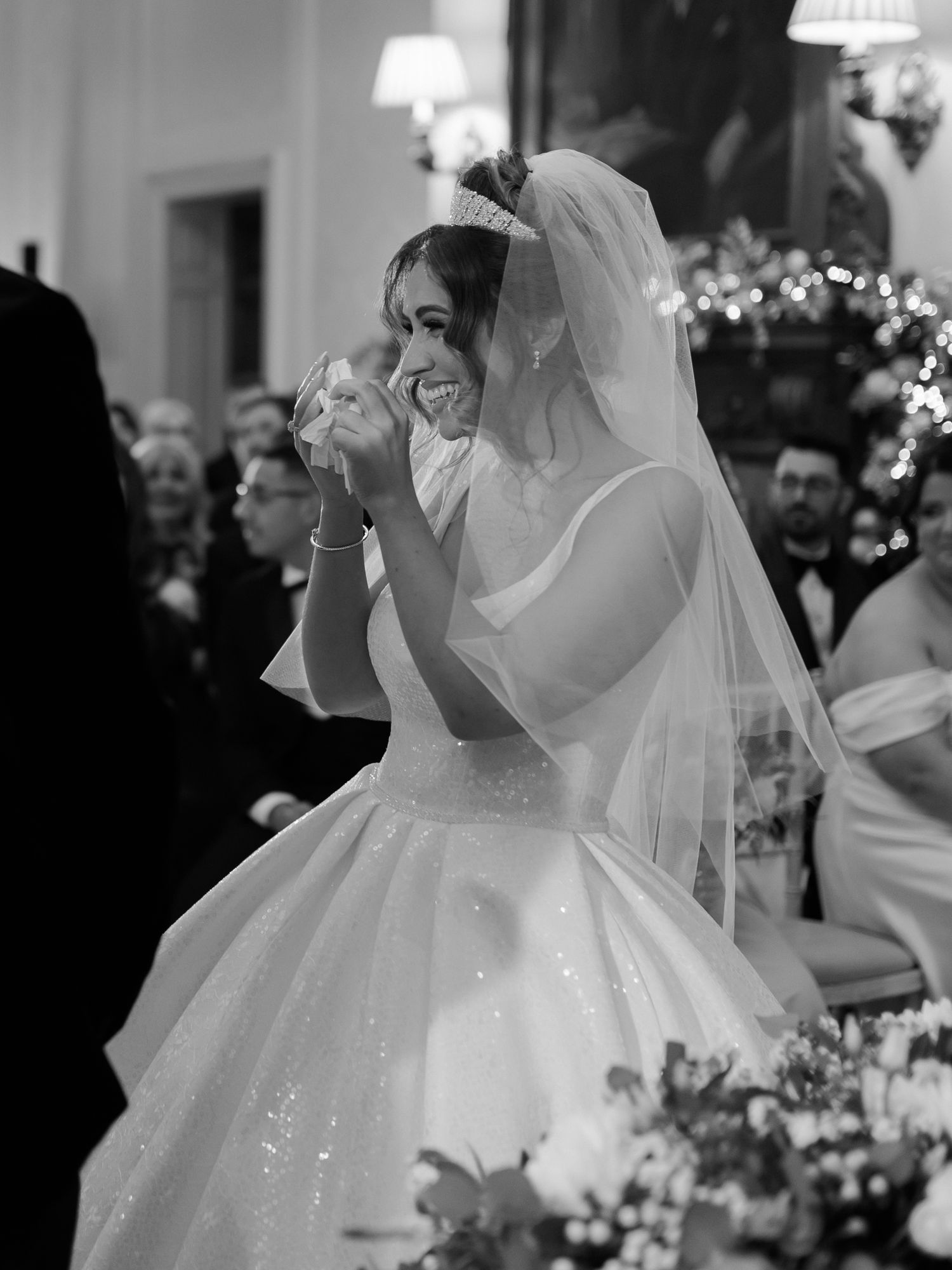 A joyful bride in a white ball gown and veil laughs during her wedding ceremony among floral decorations and guests.