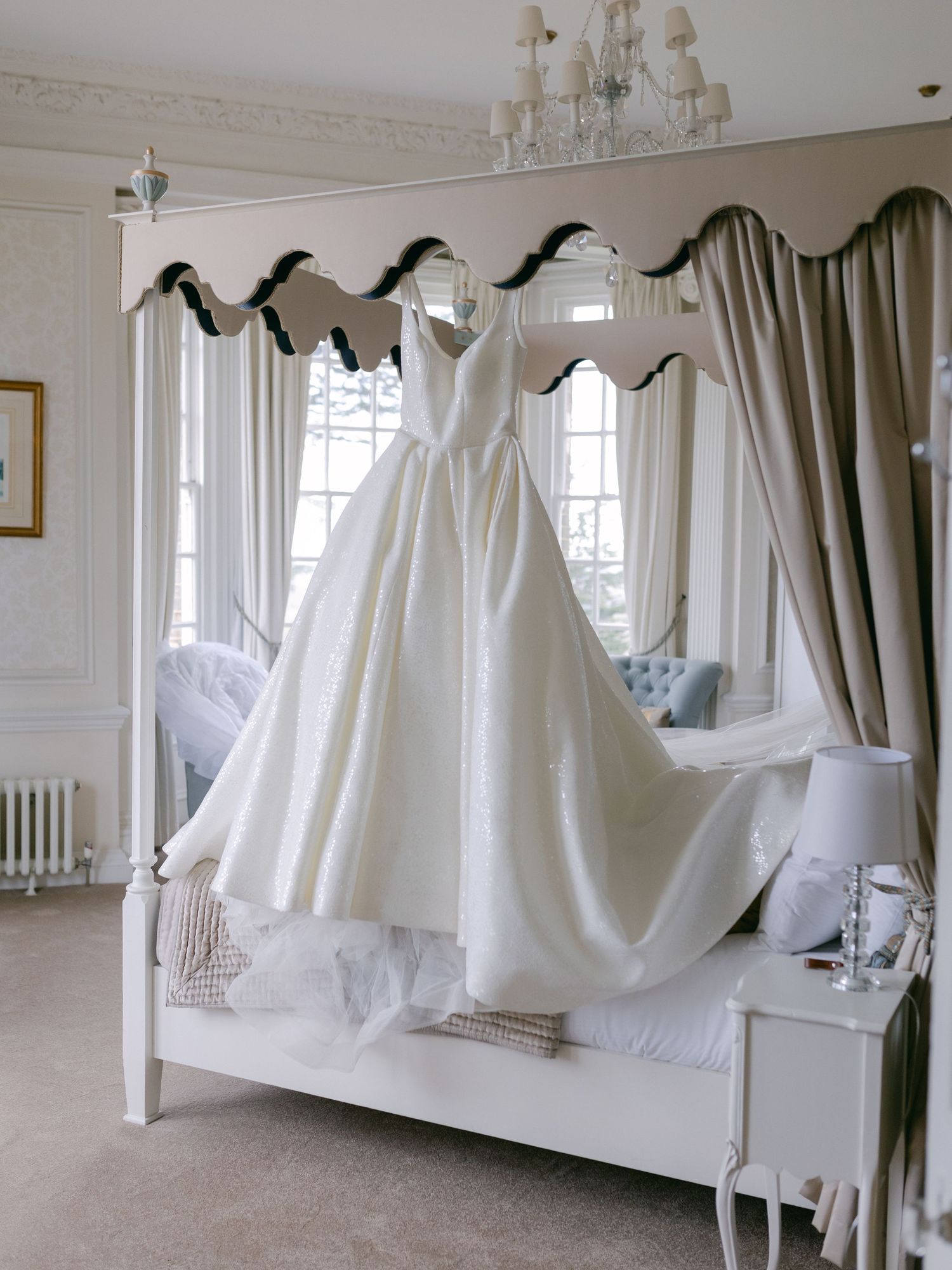 An elegant white wedding dress hangs on display over a plush bed with draped curtains and a crystal chandelier overhead.