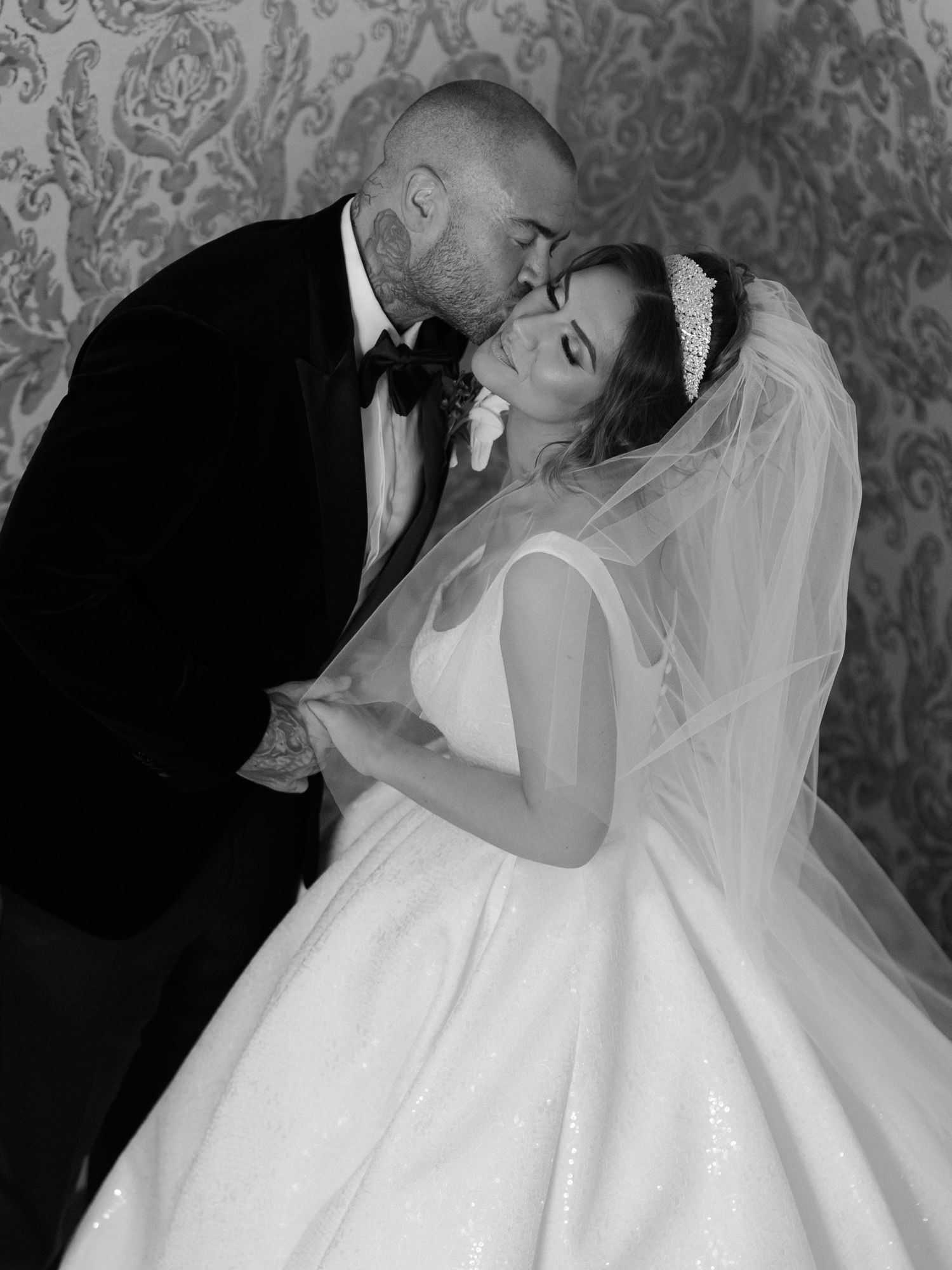 Black and white wedding photo showing romantic moment between bride in flowing gown and groom in tuxedo against patterned wall.