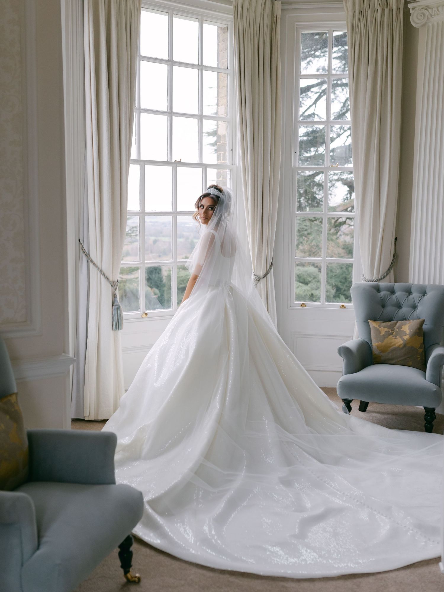 Elegant white wedding gown with long cathedral train photographed in a bright sunlit room with blue velvet chairs.