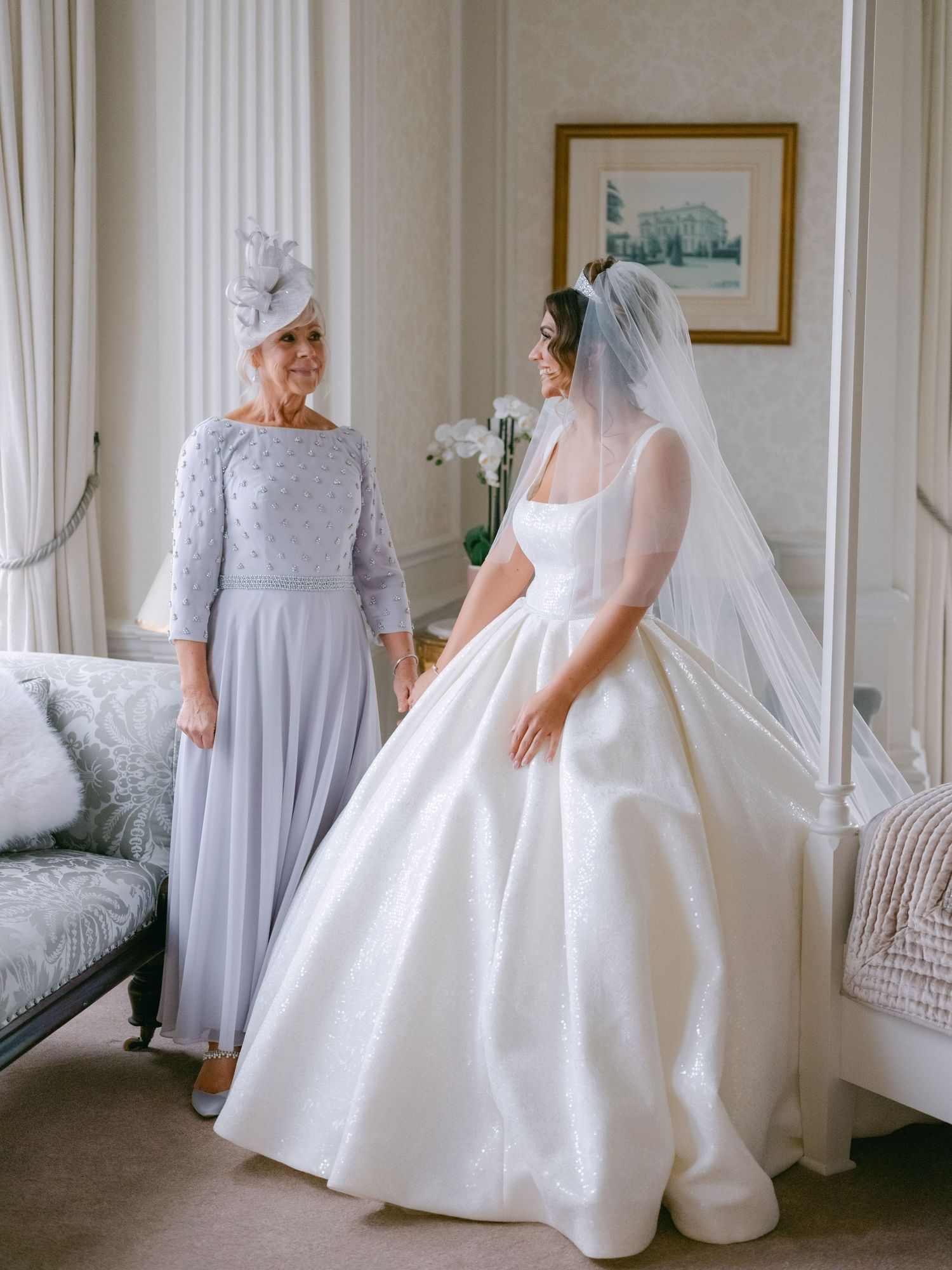 Two elegantly dressed women in a bright room with white decor, one wearing a grey gown and one in a classic white ballgown.