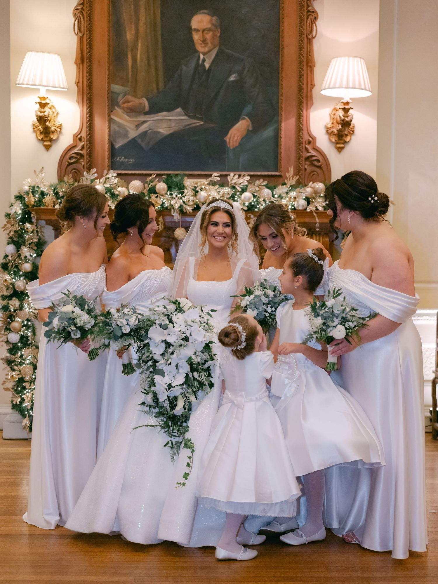 Wedding party in elegant white gowns pose together beneath ornate portrait in formal room with floral decorations.