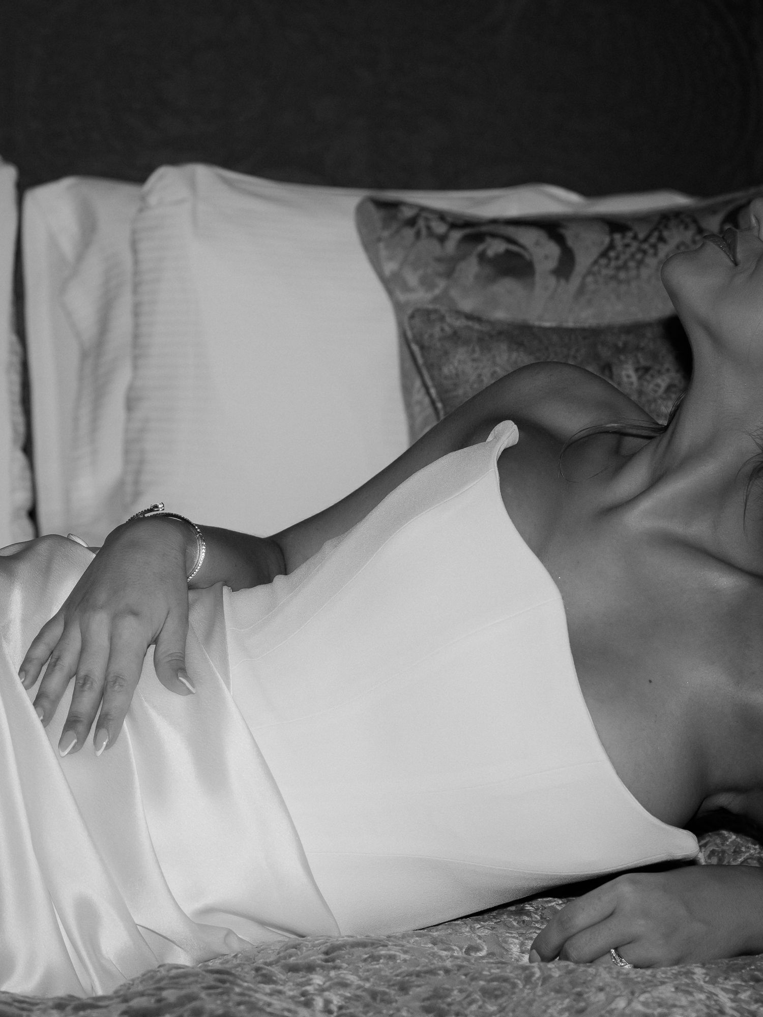Black and white intimate portrait showing someone lying on bedding wearing white satin lingerie.