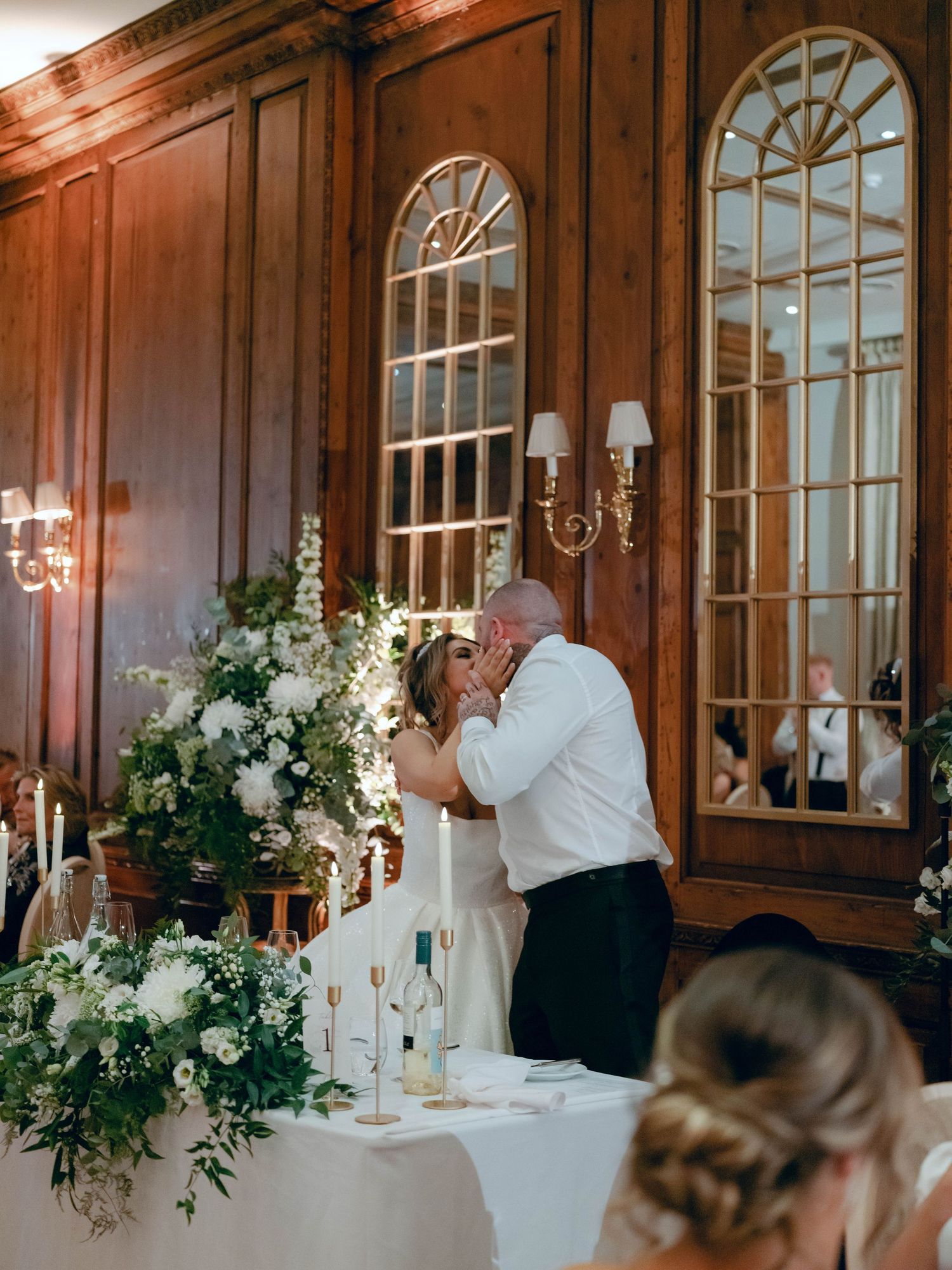 A couple shares a romantic moment at an elegant reception with arched windows, candlelight and lush floral arrangements.