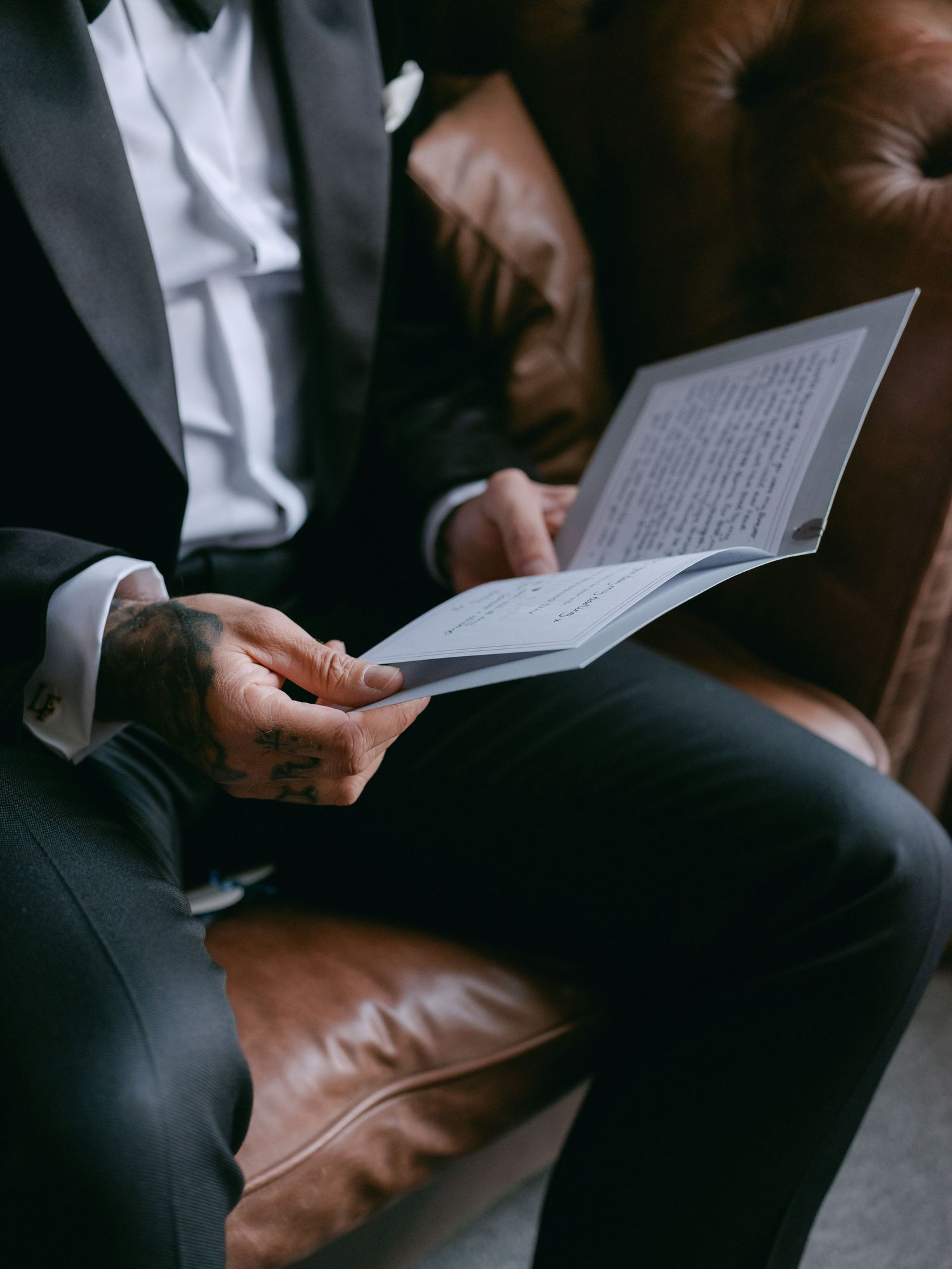 A person in formal attire sits on a brown leather chair reading a document or book.