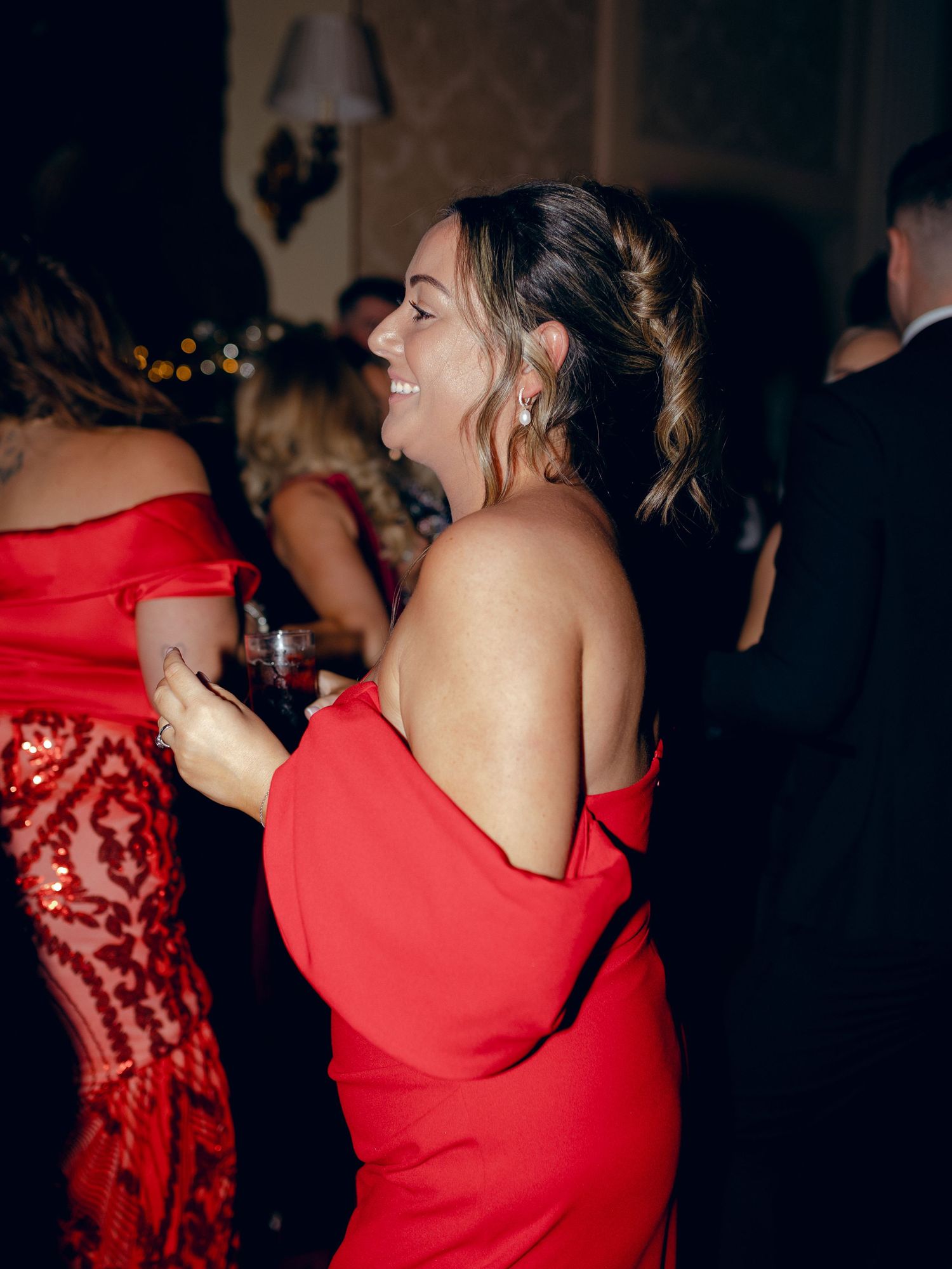 A person in a red off-shoulder dress laughs joyfully while holding a drink at an evening party.