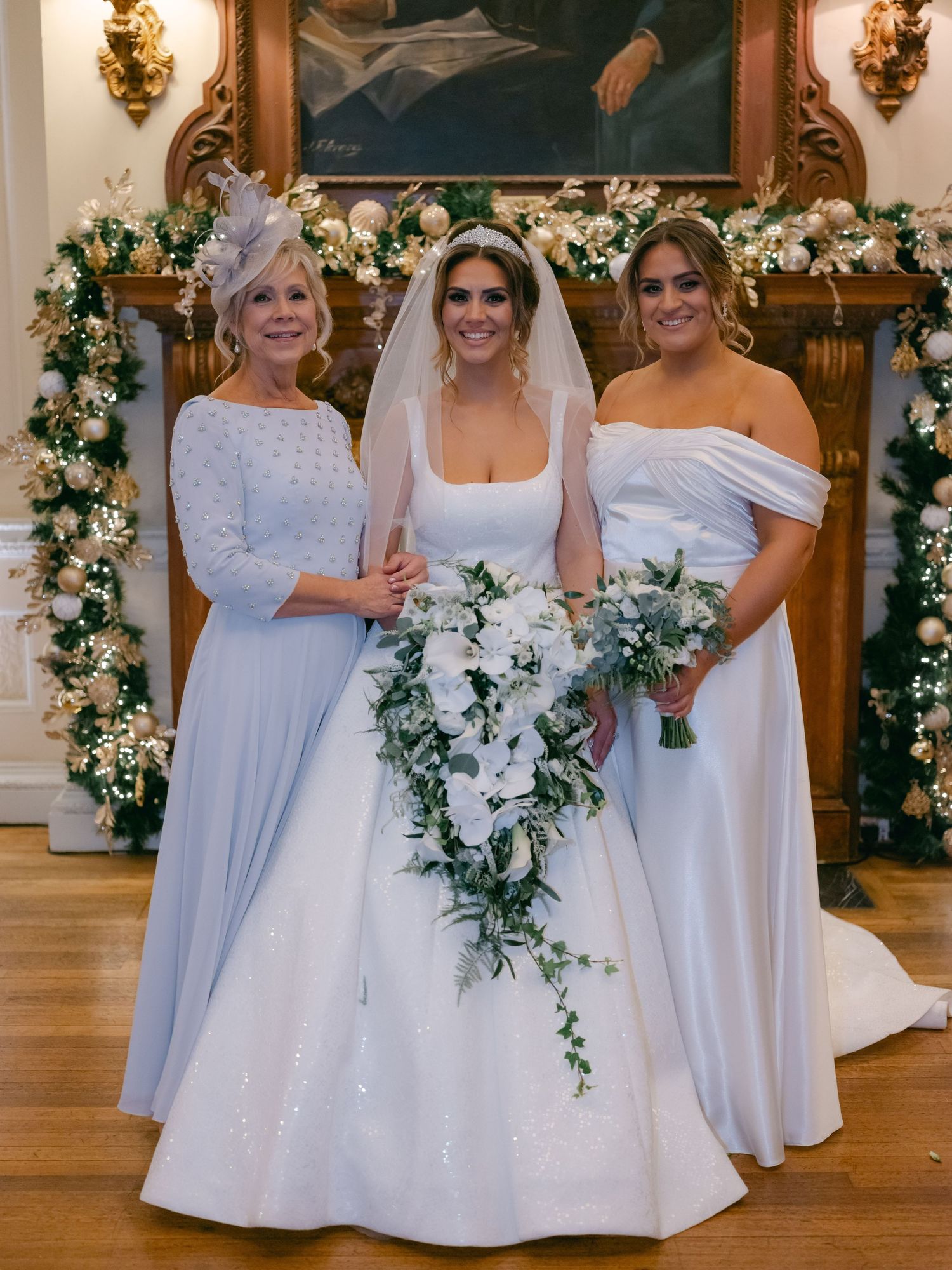 Three women stand together at a wedding ceremony with floral arch decorations in an elegant indoor setting.