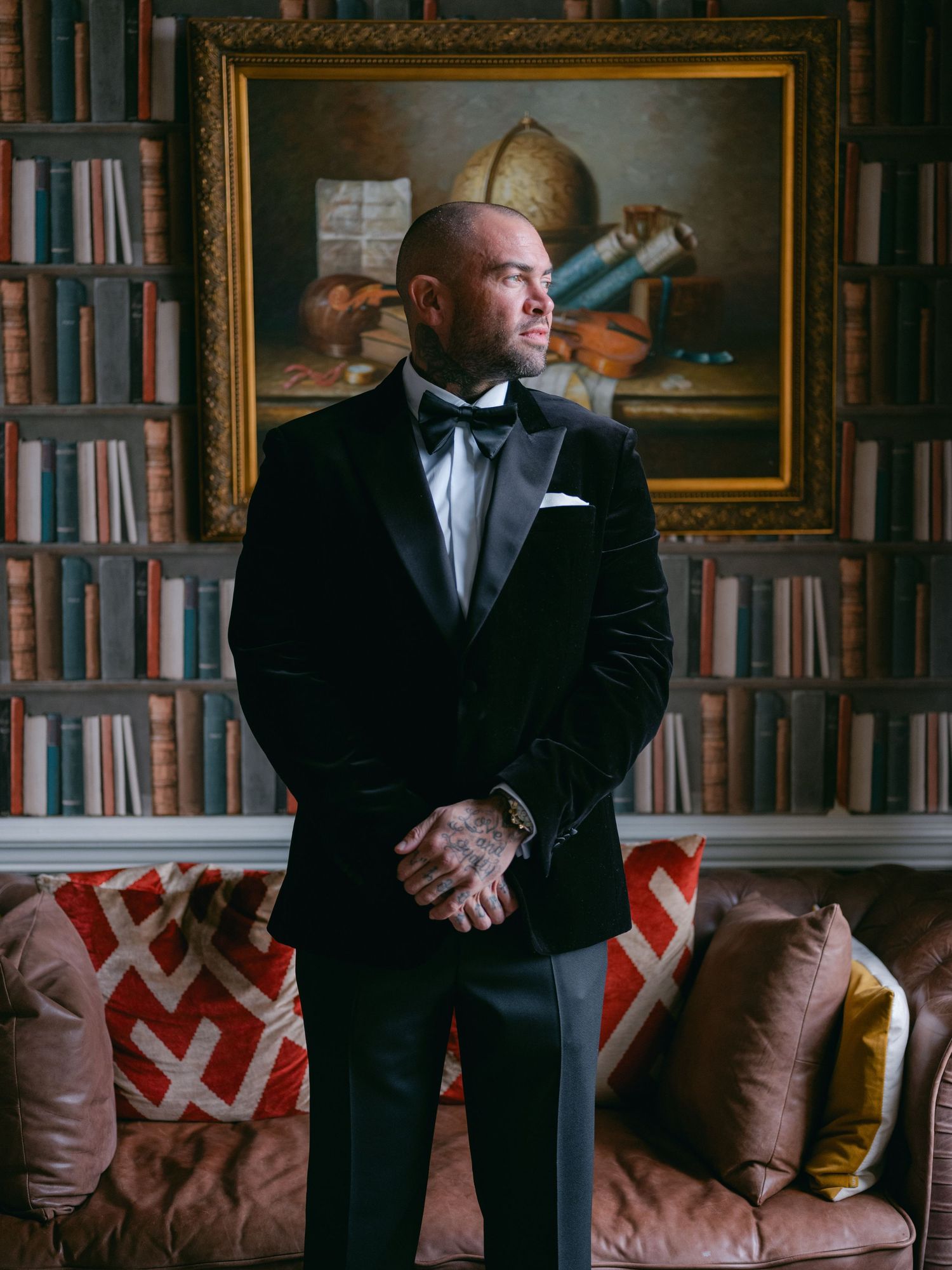 Elegant formal portrait in library setting shows person wearing black tuxedo and bowtie against backdrop of bookshelves and artwork.