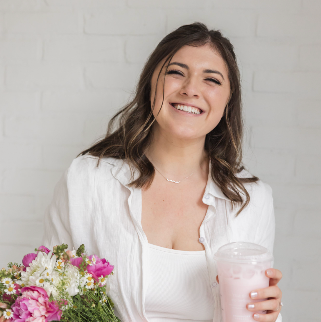 A smiling person in white holds a pink beverage while standing next to fresh flowers against a light background.