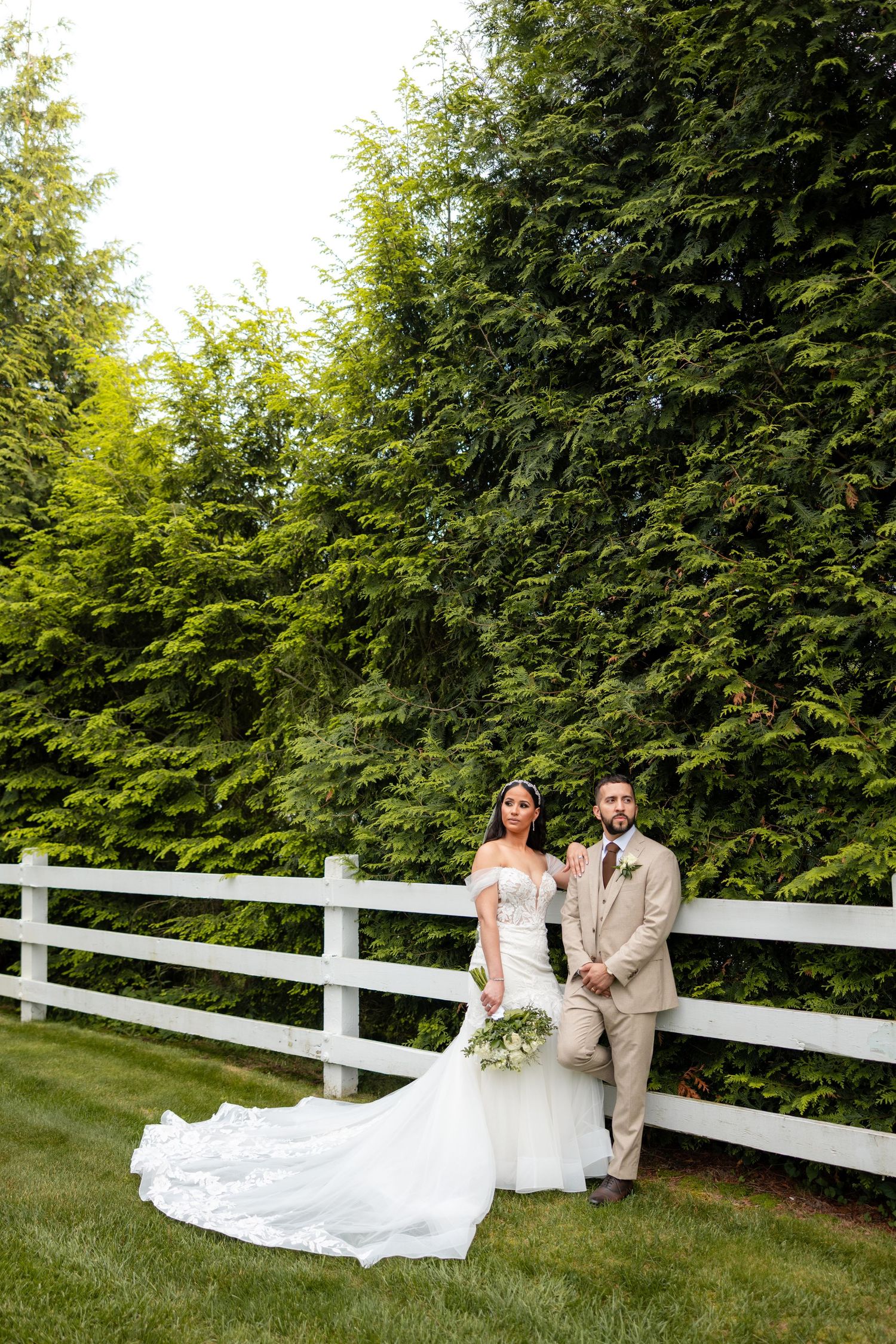 Wedding couple poses for a romantic photo series next to a white fence surrounded by lush green trees.