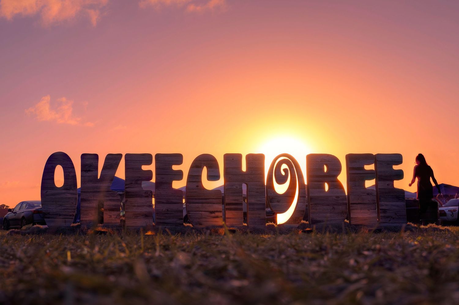 Giant Okeechobee letters silhouetted against a vibrant orange sunset sky with palm trees in the background.