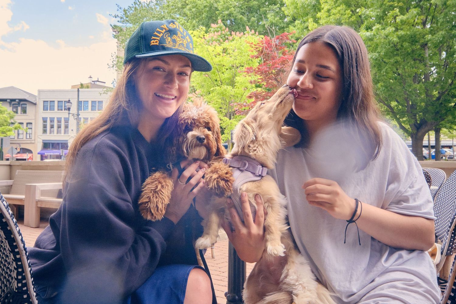 Two people share a joyful moment holding a small brown puppy outdoors on a sunny day near some trees and buildings.