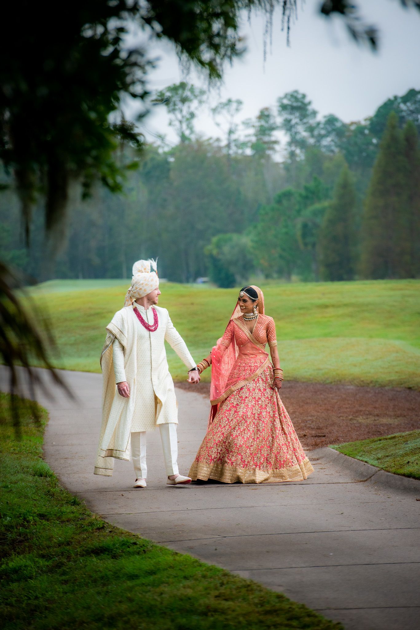 Couple in traditional Indian wedding attire walking together on a path surrounded by trees.
