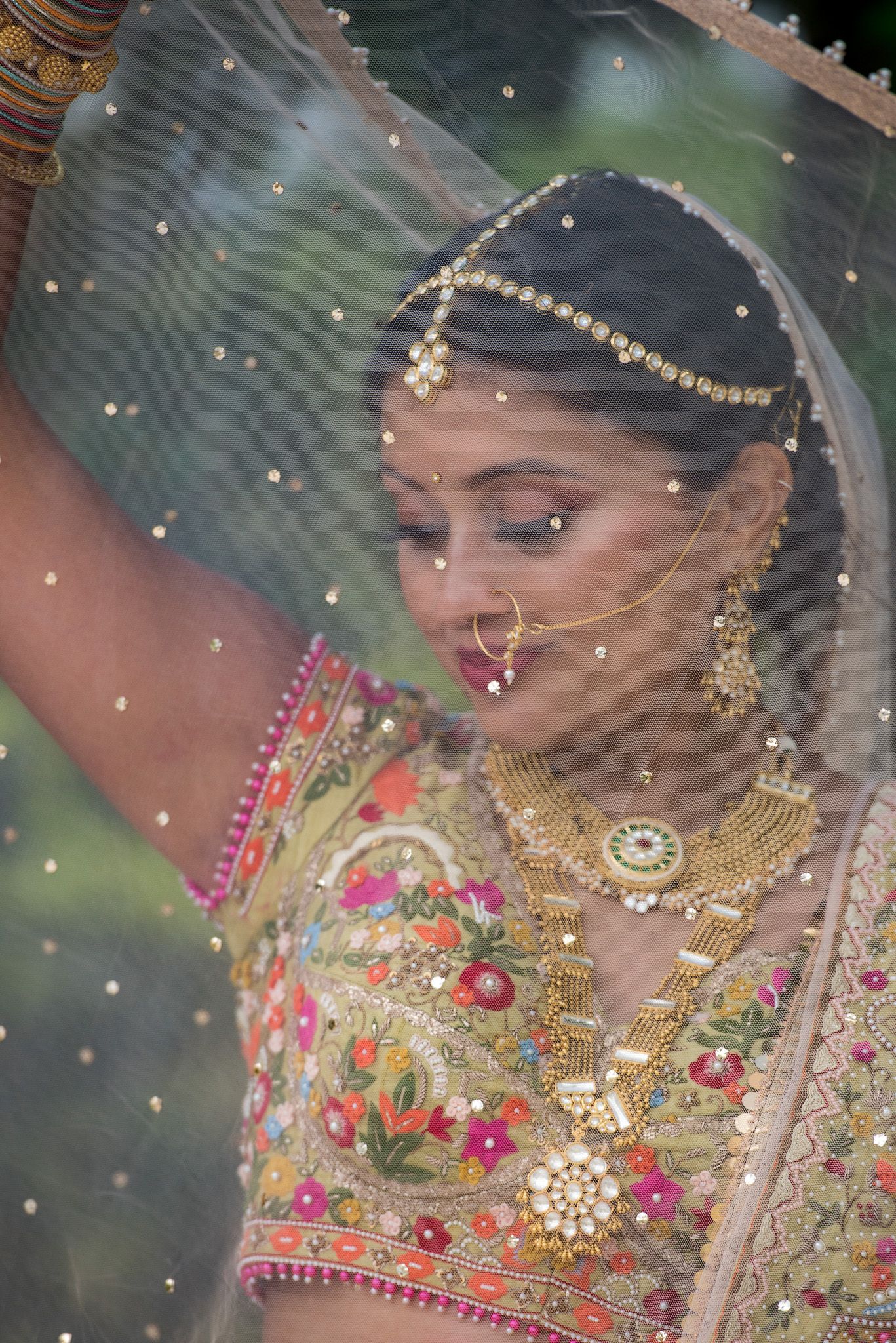 Traditional Indian bride in ornate jewelry and embroidered attire under a sheer veil.
