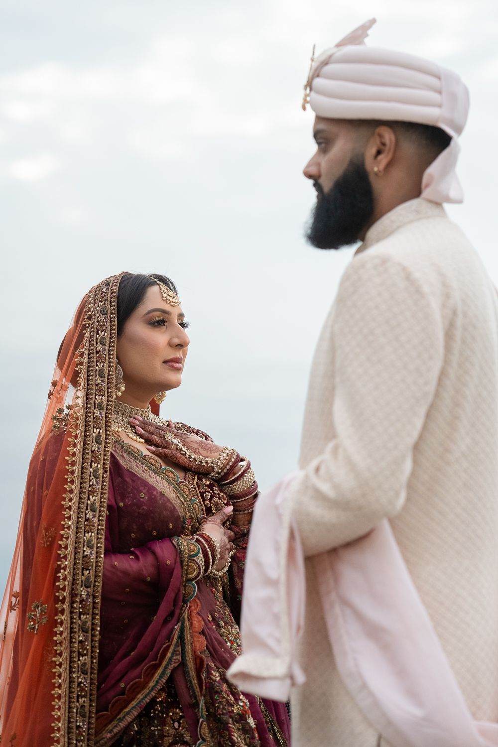 Traditional South Asian wedding couple in ornate ceremonial attire against cloudy sky.