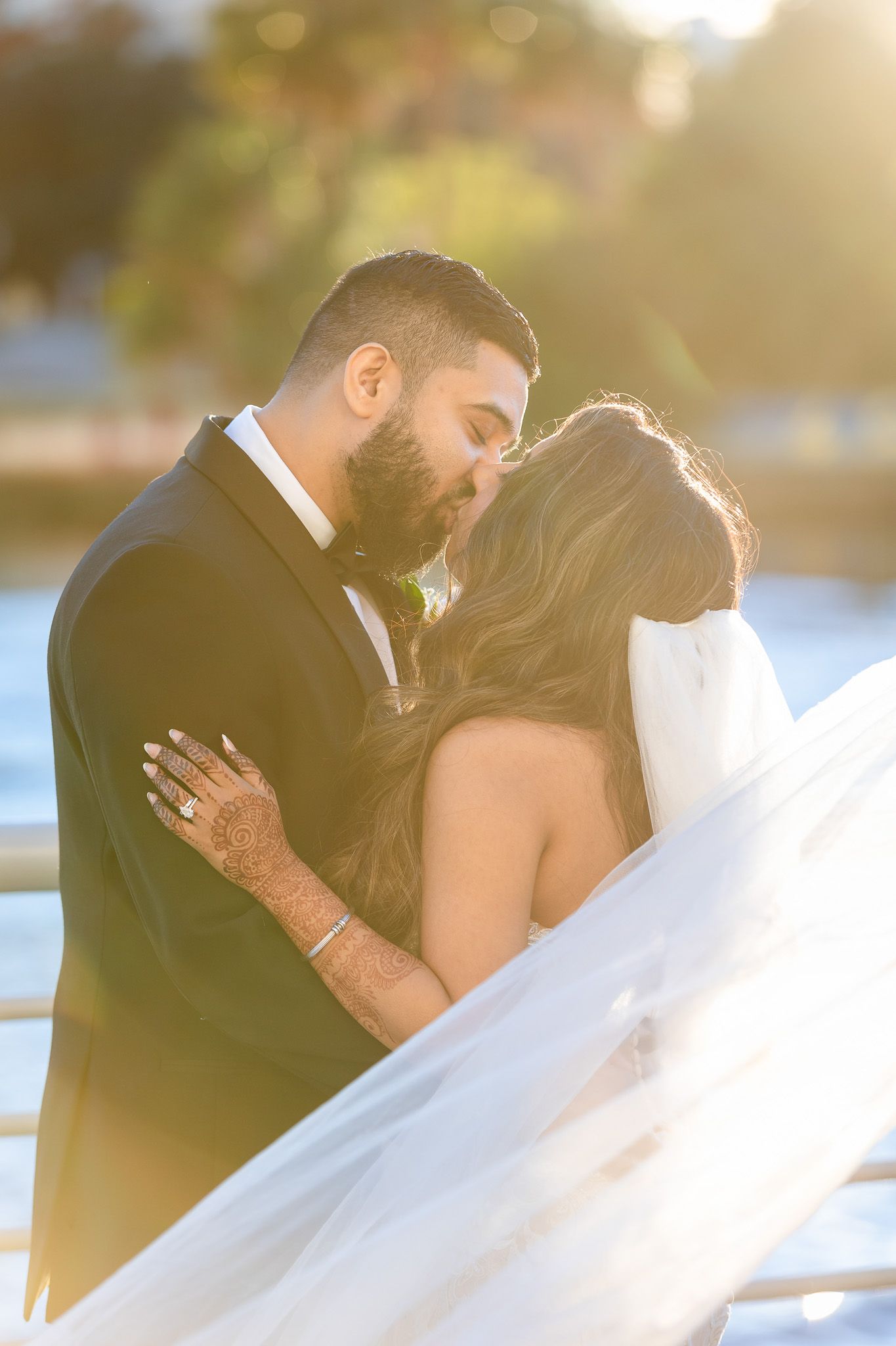 Romantic sunset wedding portrait with flowing veil and waterfront backdrop.