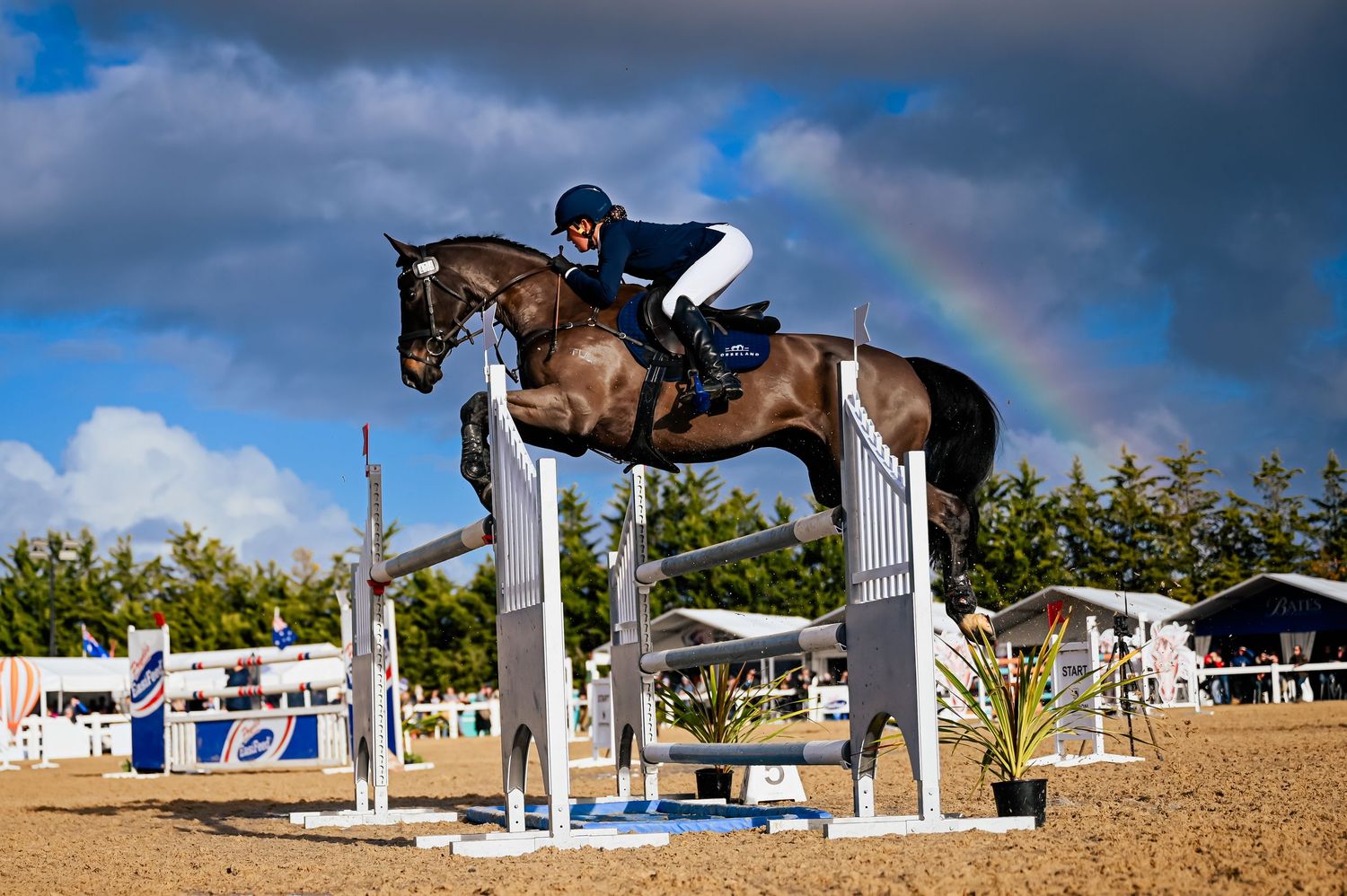 Horse and rider jumping over white poles during competition with dramatic cloudy sky.