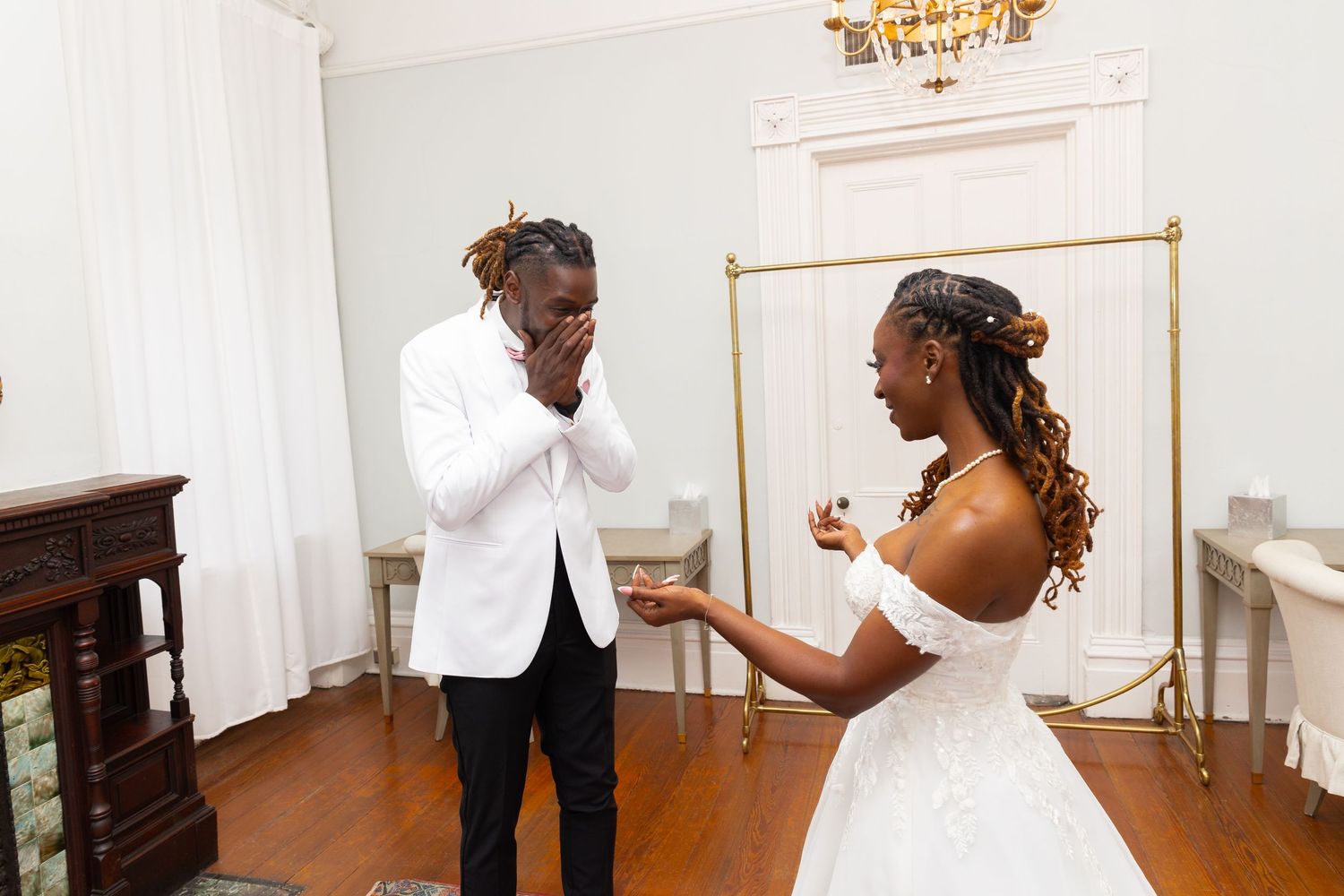 A joyful couple shares an emotional first look moment together in an elegant white room with a gold chandelier.