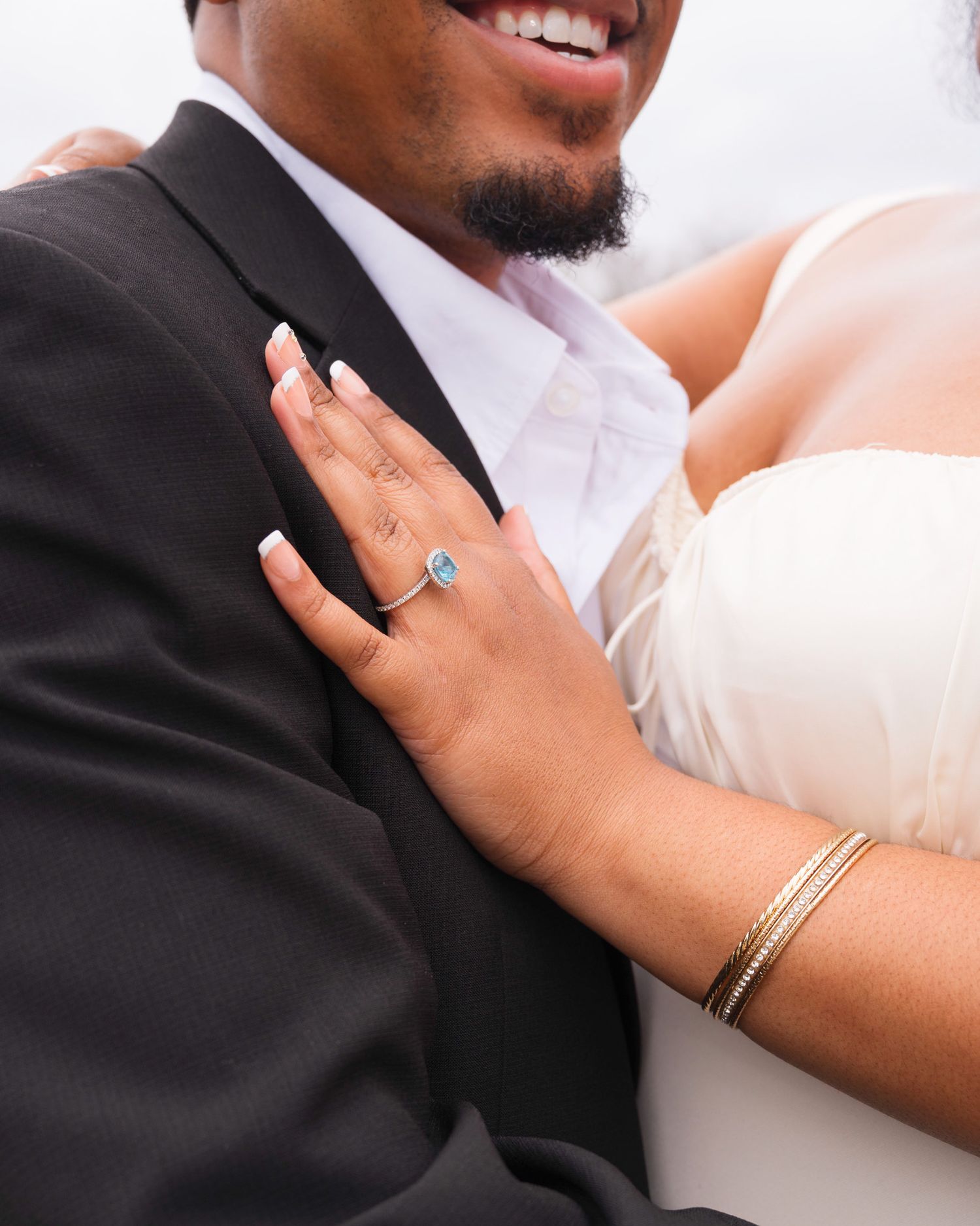 A close-up wedding photo showing hands with an engagement ring and bracelet against a black suit jacket.