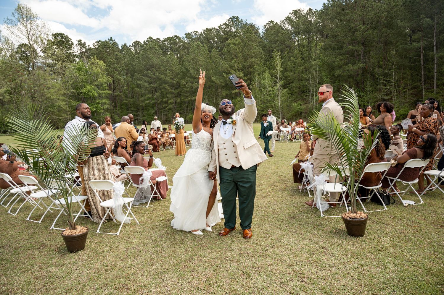 Newlyweds celebrate with raised arms while walking down the aisle at an outdoor wedding ceremony with guests seated on white chairs.