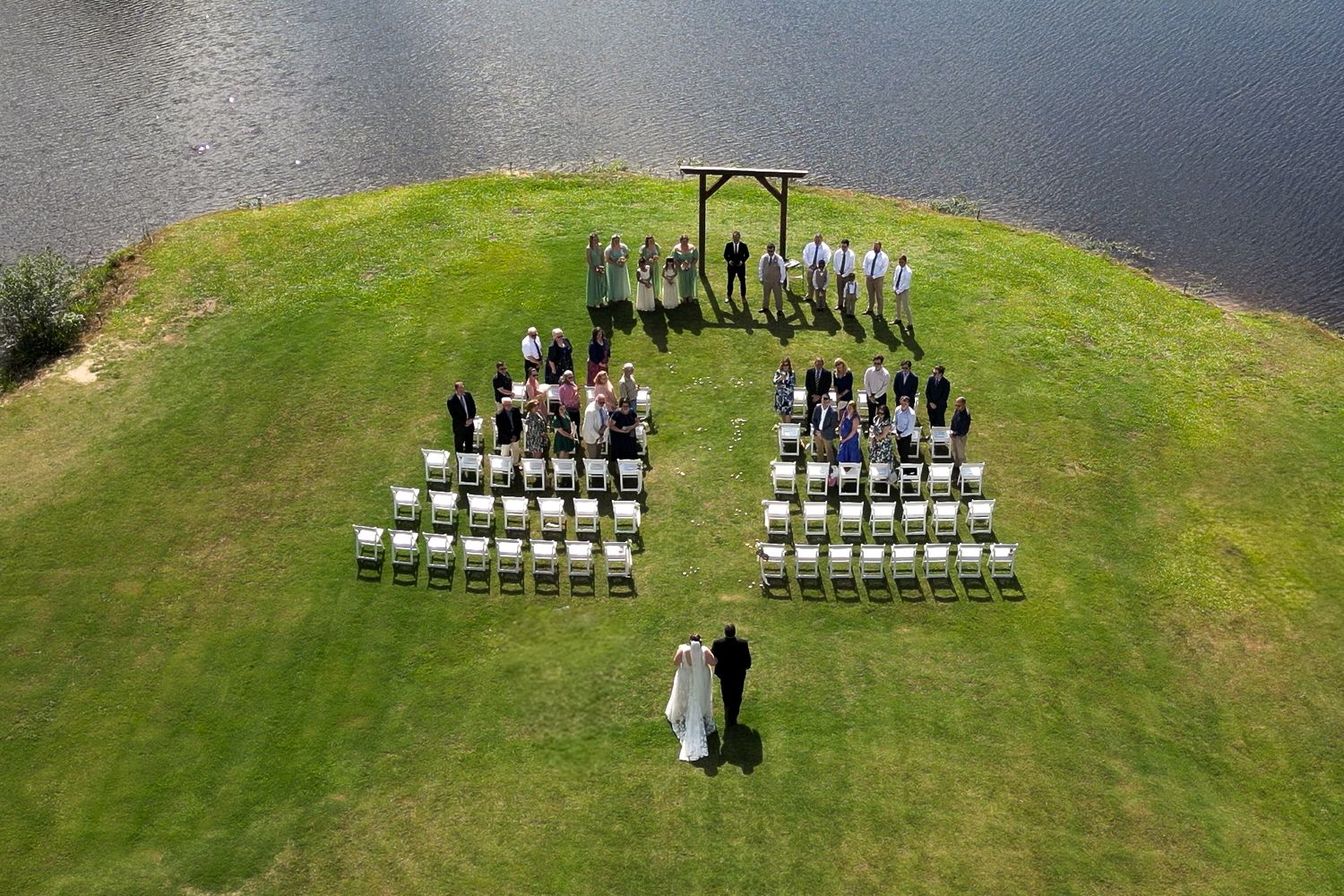 Aerial view of an outdoor wedding ceremony setup with white chairs arranged in rows on a grassy peninsula by the water.