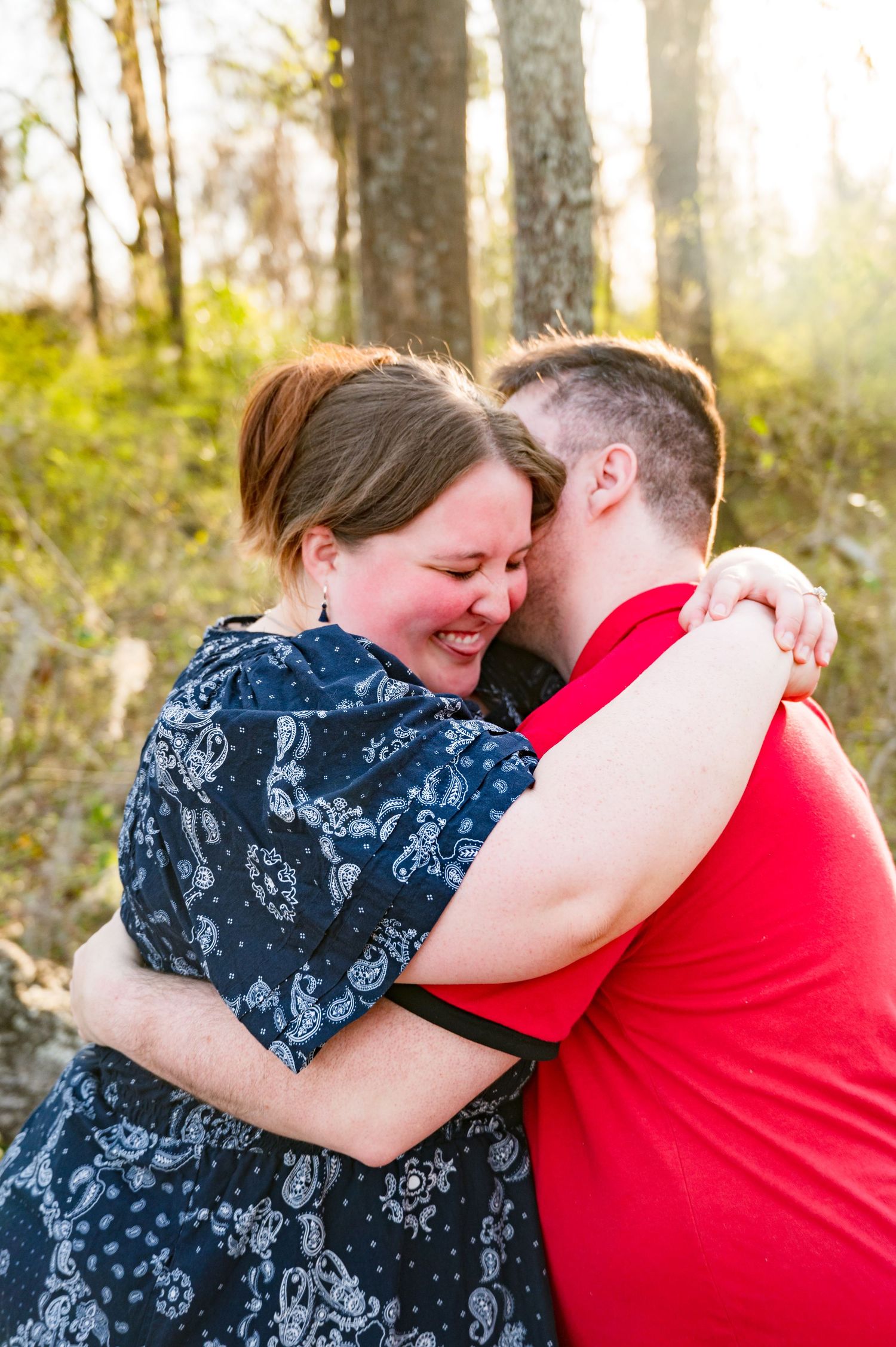 A couple embraces lovingly in a sun-dappled forest setting wearing red and blue floral outfits.
