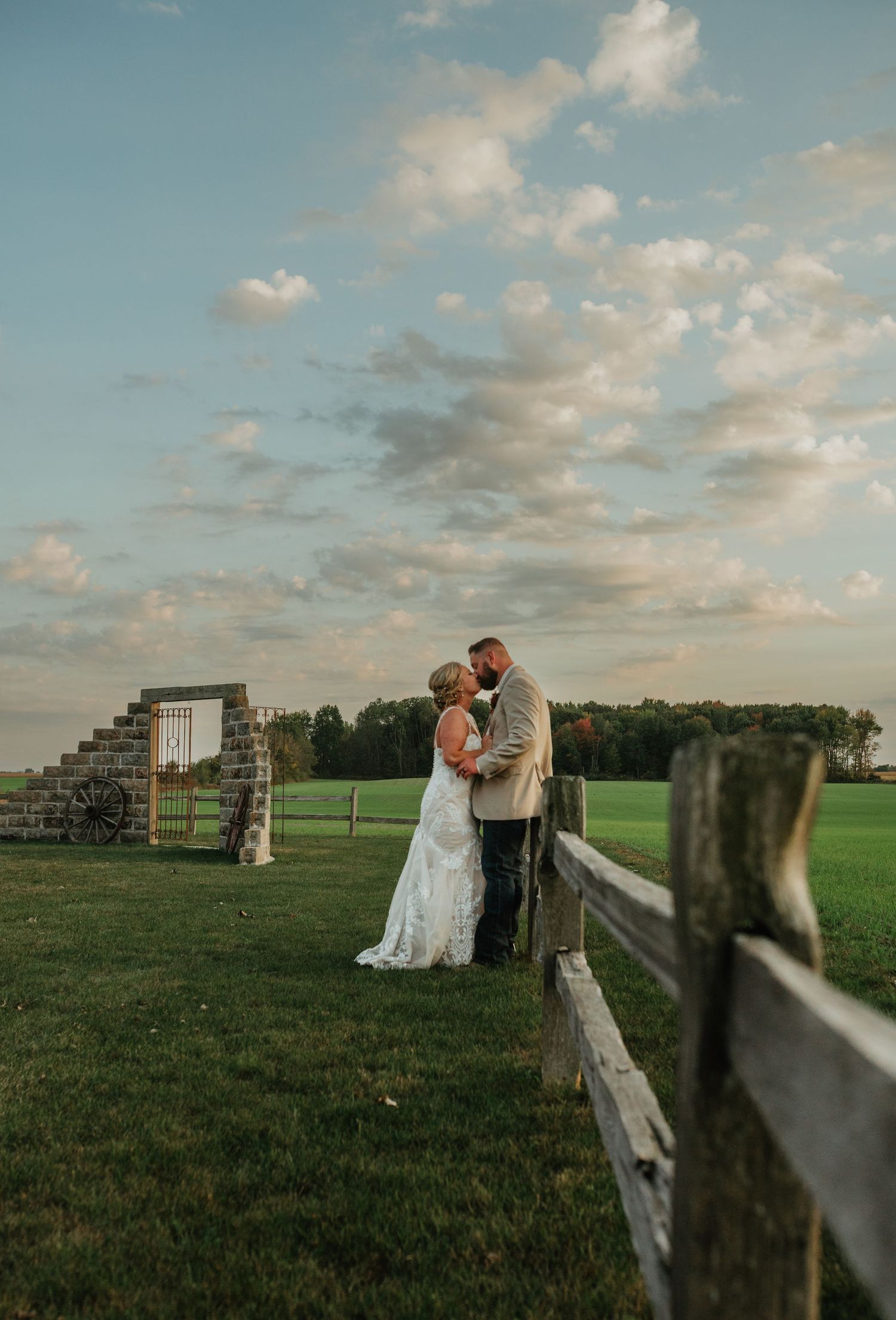 A couple shares a tender moment at sunset by a rustic wooden fence on a green countryside field.