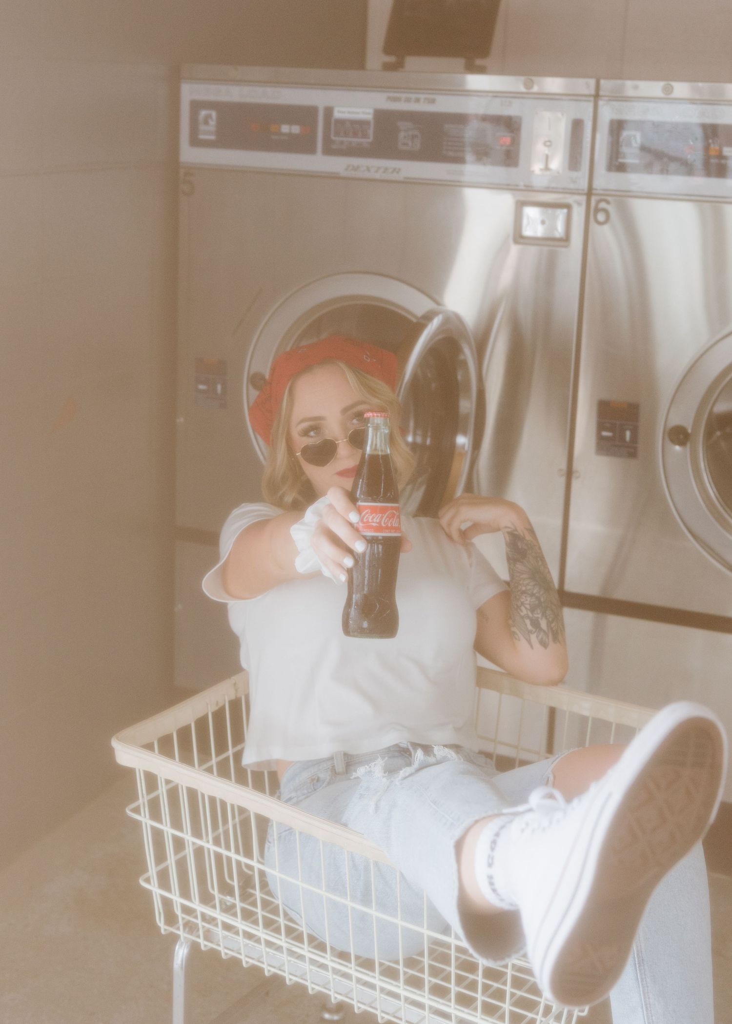 Person sitting inside white laundry basket taking mirror selfie in retro-style laundromat setting.