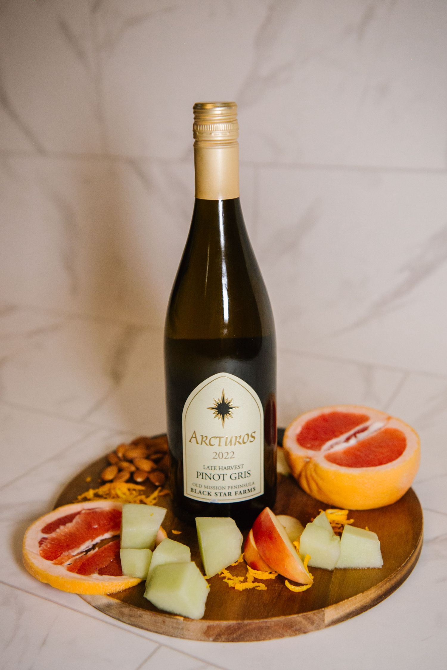 Bottle of Arcturus Pinot Gris wine on wooden board with sliced grapefruit and honeydew melon garnished with orange flowers.