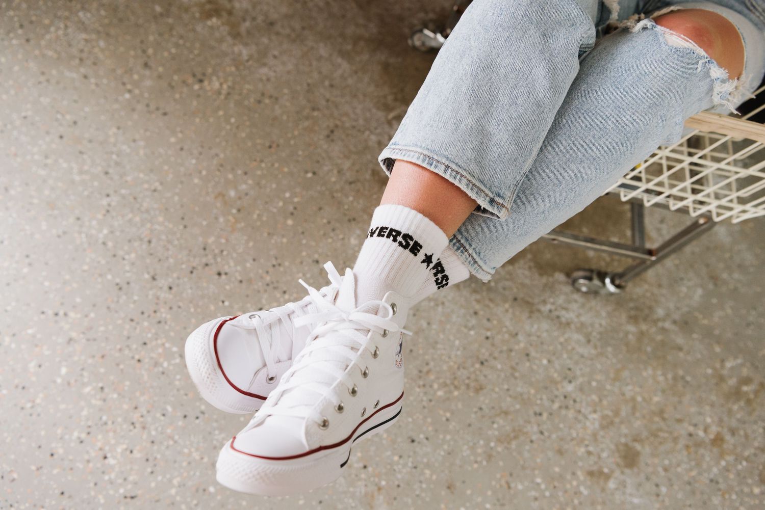 A pair of white Converse sneakers with red trim worn with light-wash jeans and black text socks on a concrete floor.