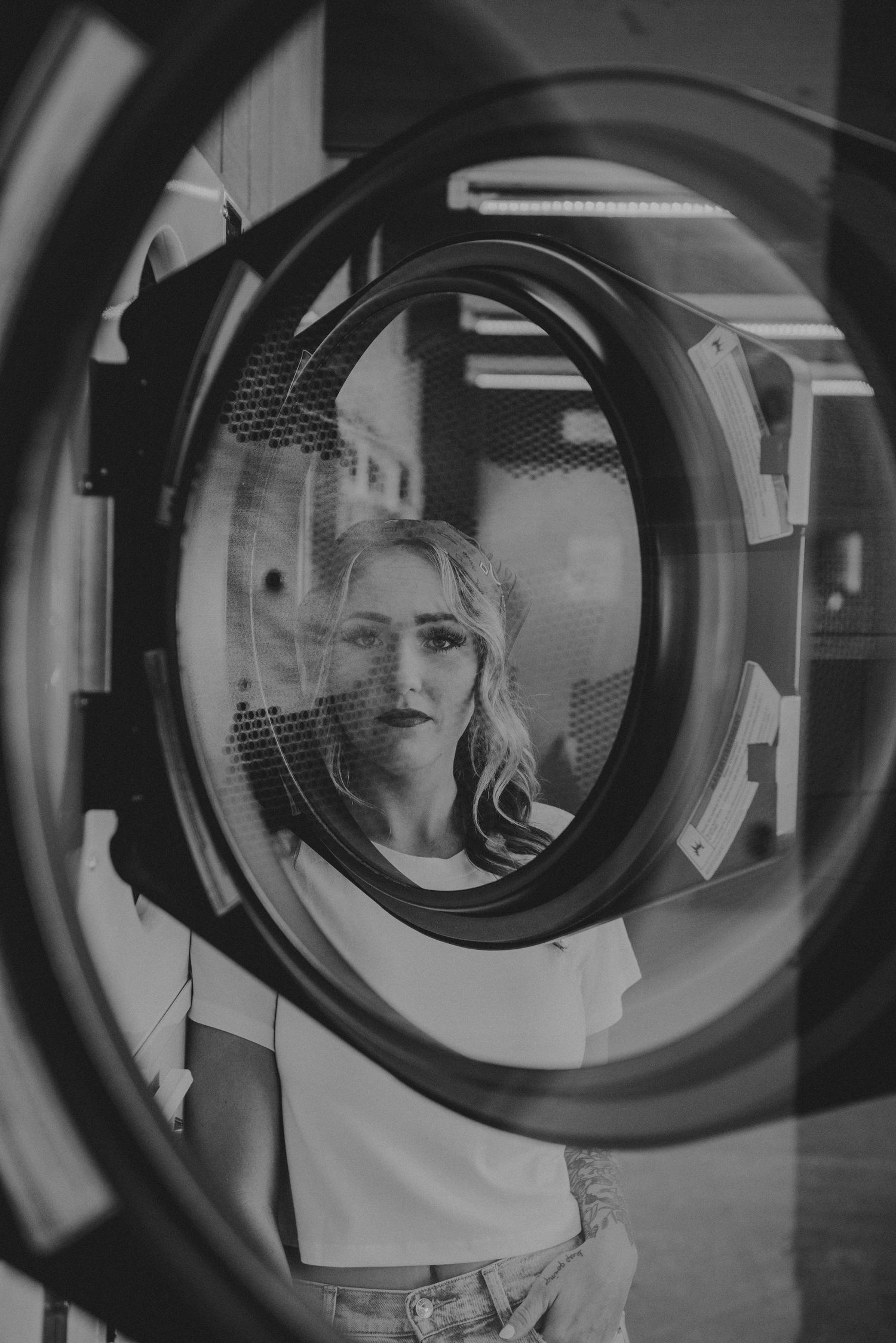 Artistic black and white photo of someone viewed through circular washing machine door window at laundromat.