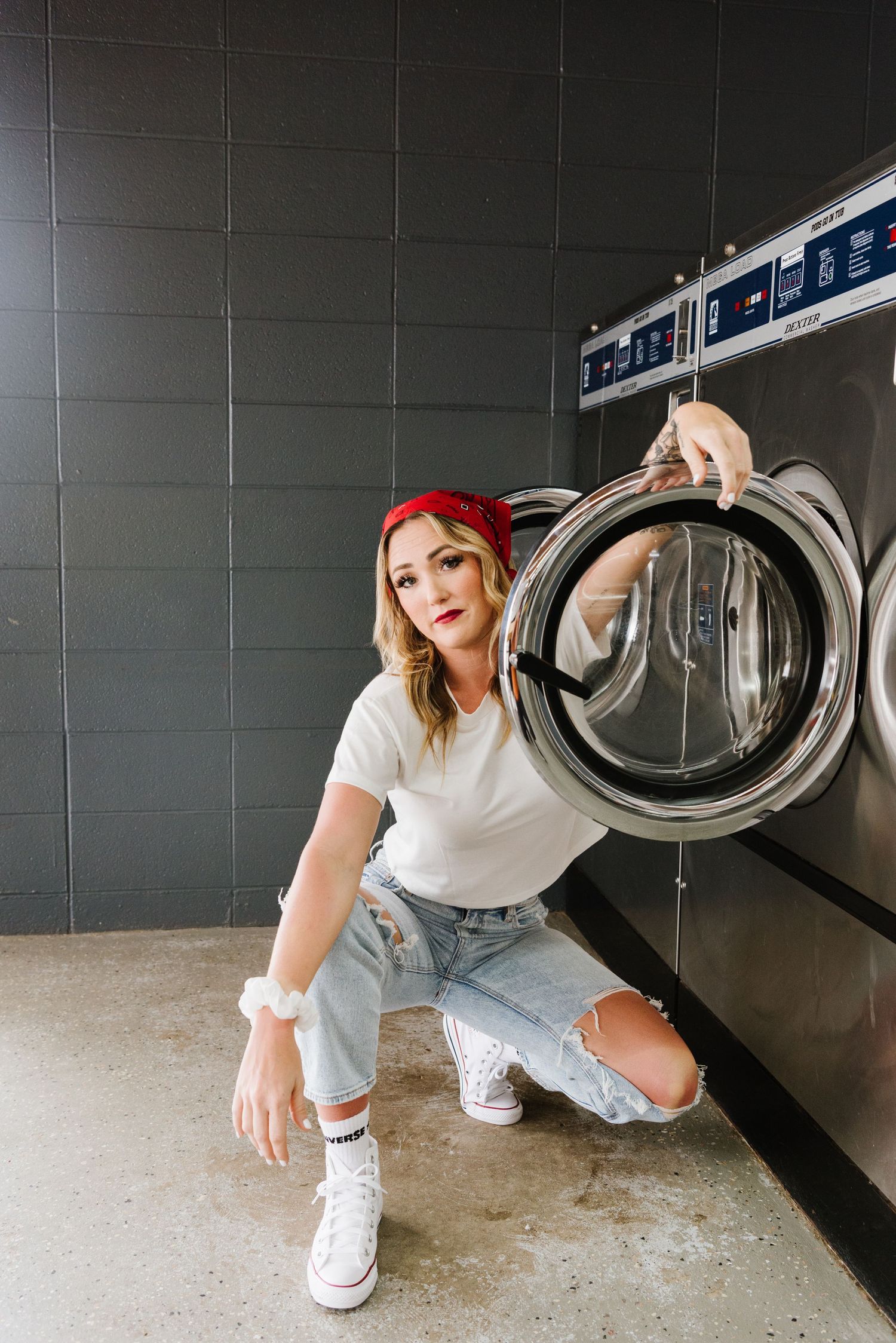 Person in ripped jeans and red bandana poses at laundromat with washing machine door open.