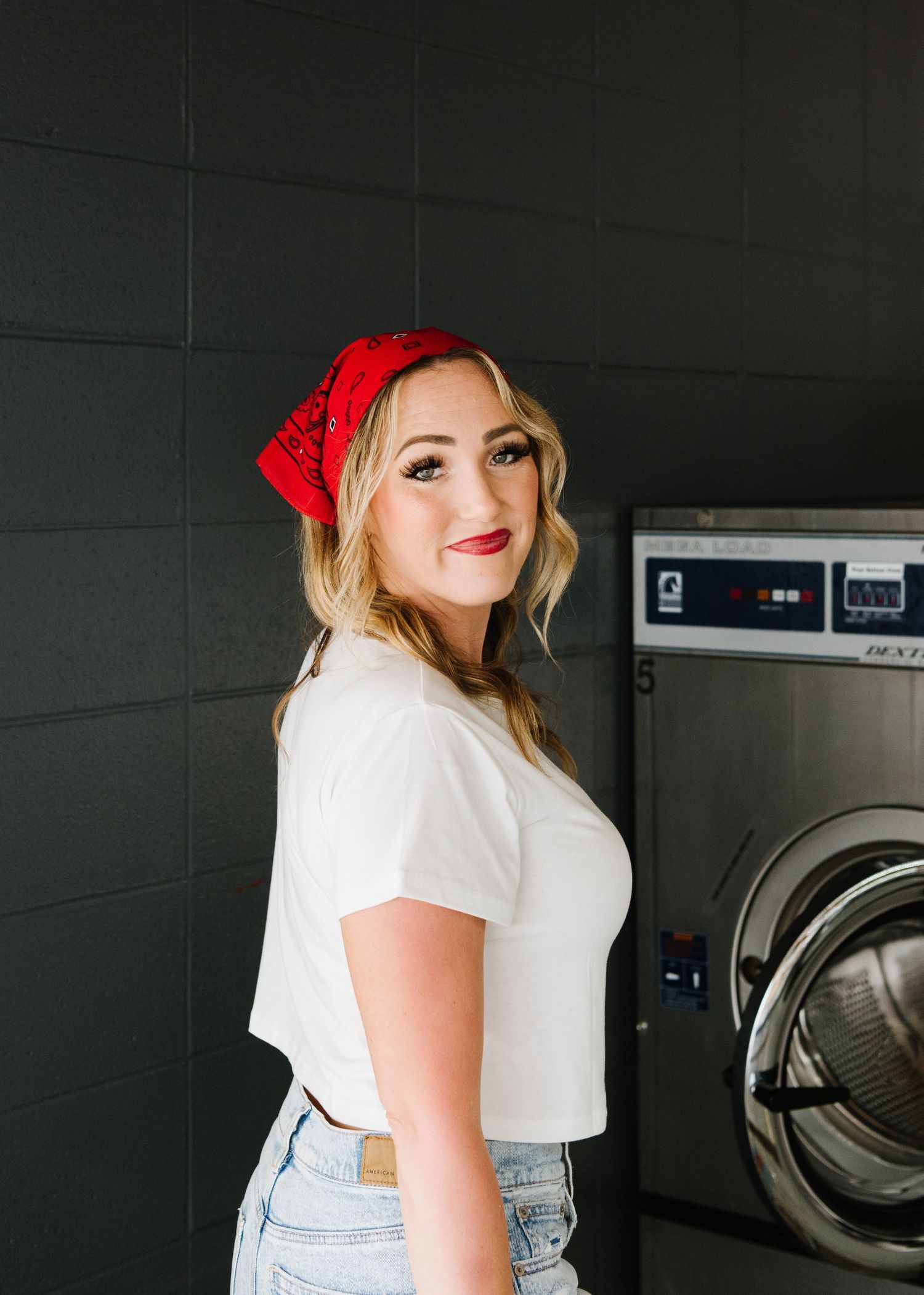 Someone wearing a red bandana and white t-shirt poses at a laundromat next to commercial washing machines.