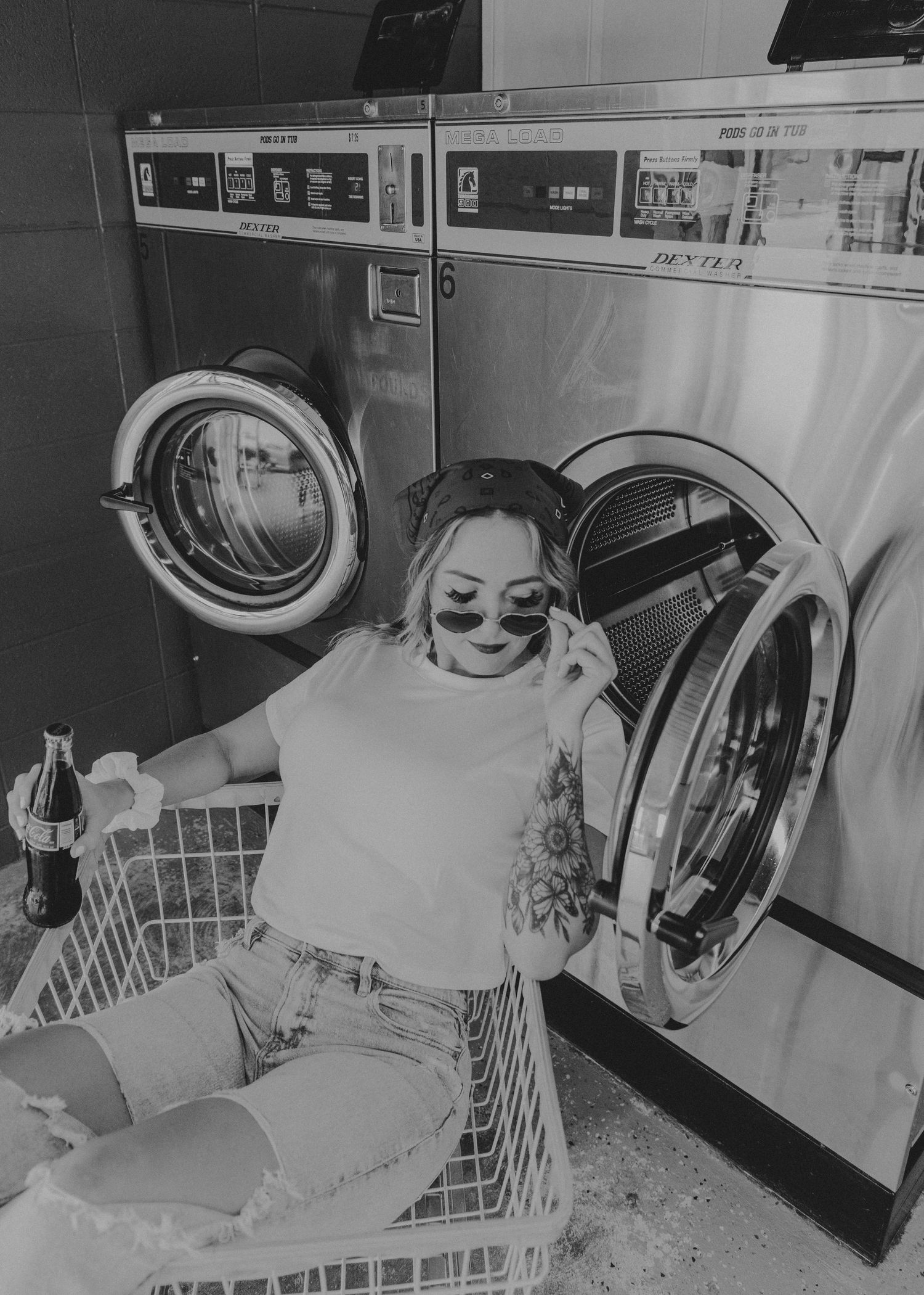 A person sits smiling in a laundromat cart next to a washing machine in a black and white photograph.