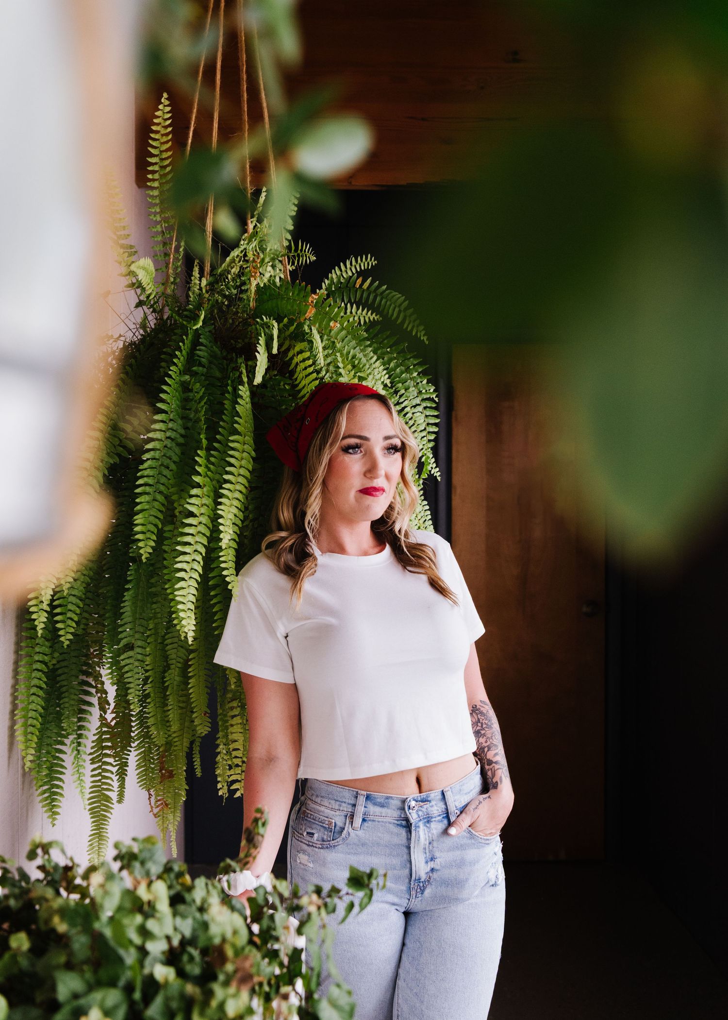 Indoor portrait with hanging fern plant and casual outfit of white crop top and blue jeans against dark background.