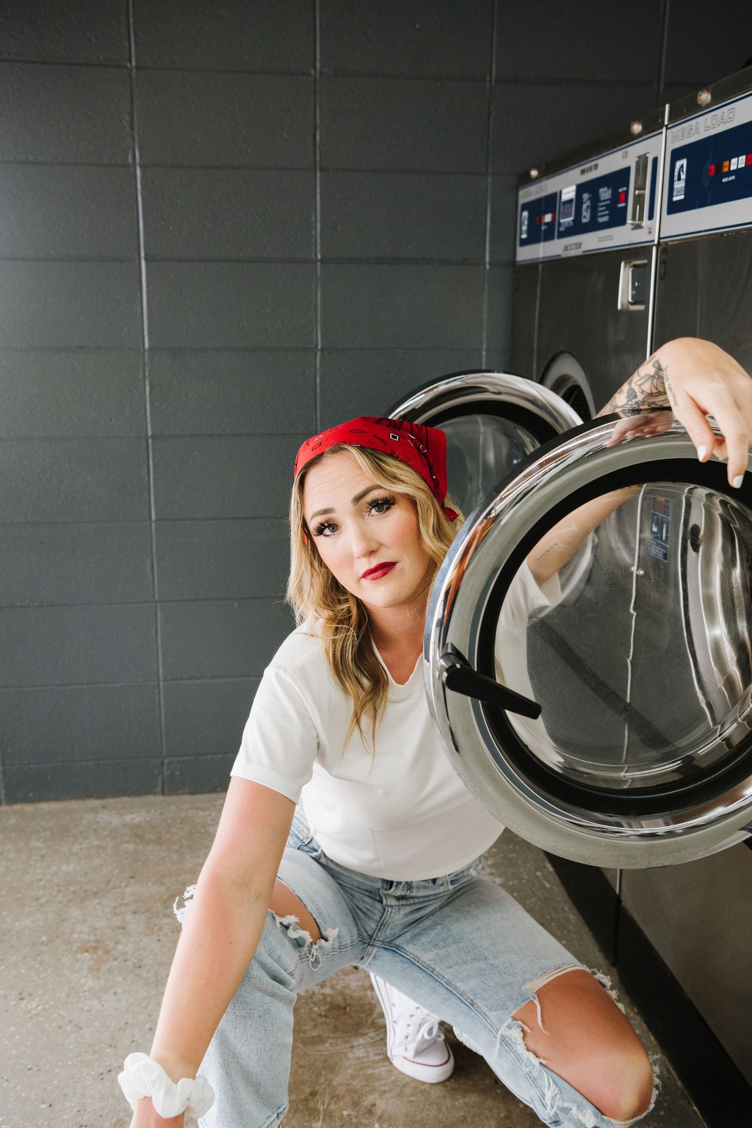 Woman in red bandana and ripped jeans poses in front of industrial washing machine at laundromat.