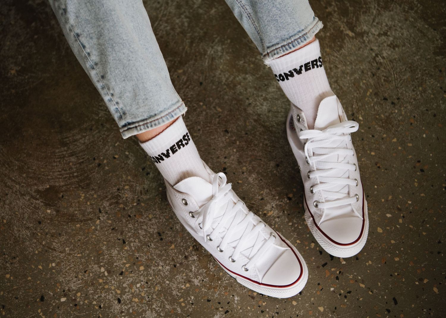 Person wearing light blue jeans and white Converse All Star sneakers with branded socks on concrete surface.