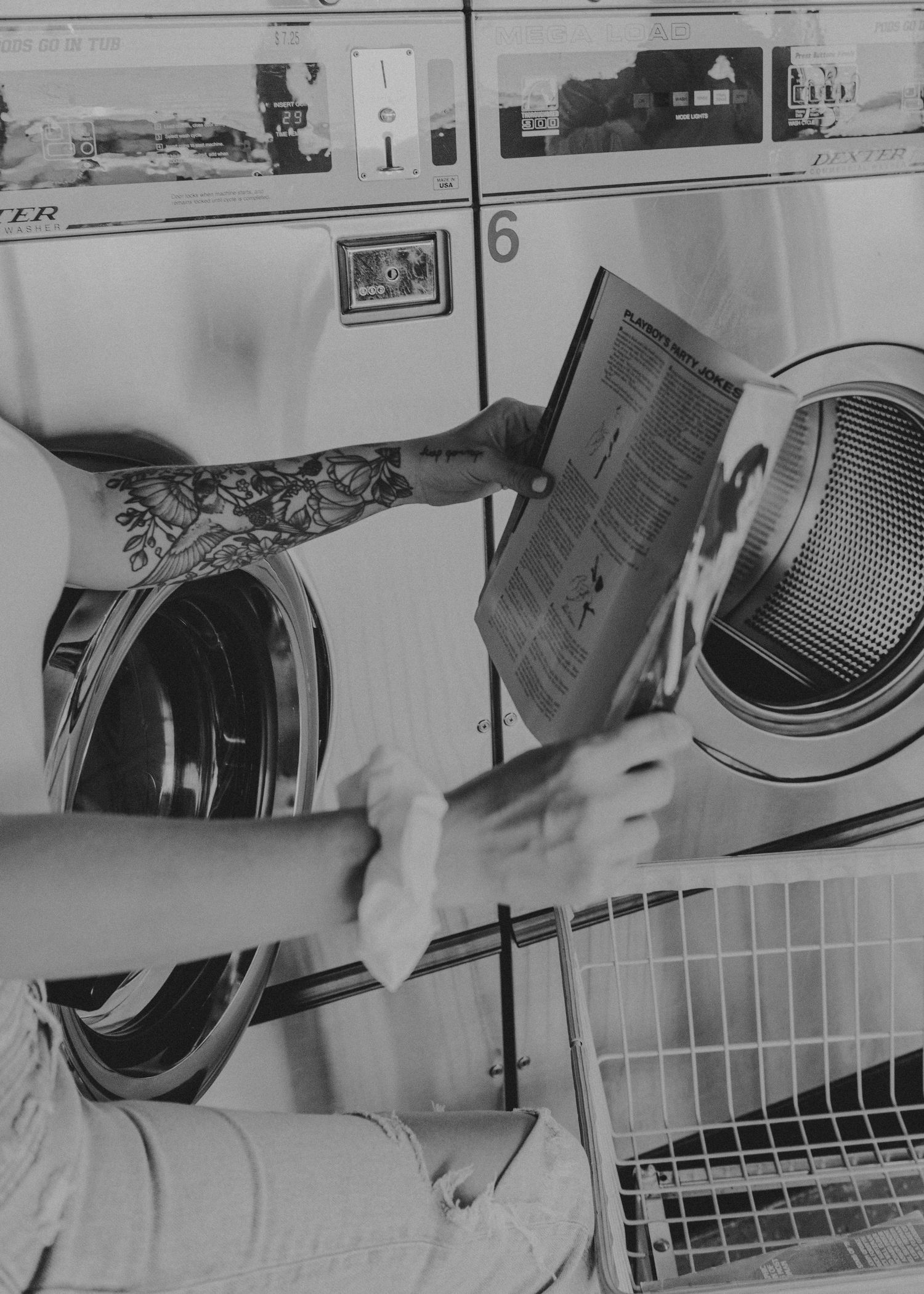 Someone reads a magazine while waiting at a laundromat with front-loading washing machines visible in black and white.