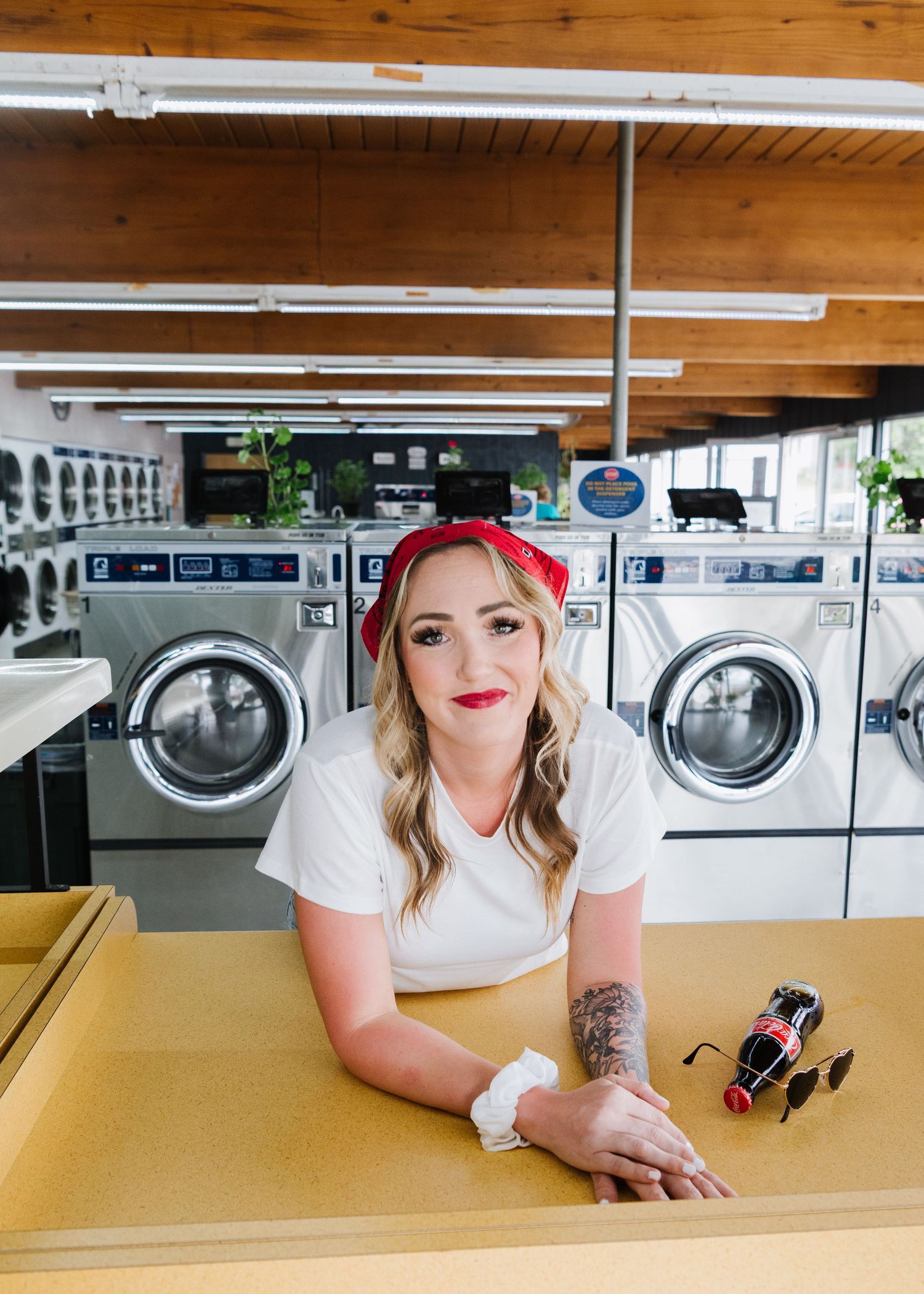 Interior view of a laundromat with rows of washing machines and someone sitting at a yellow table wearing a red bandana.
