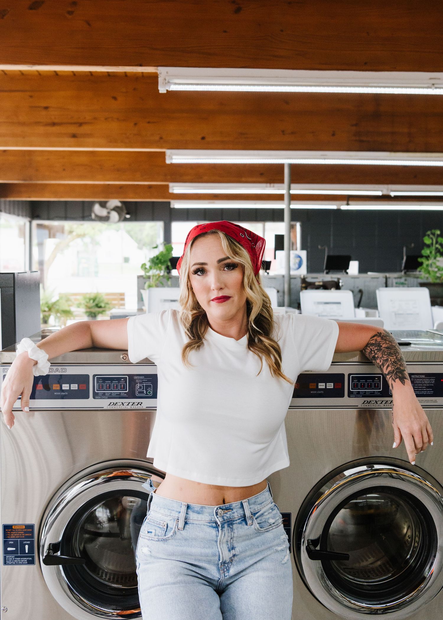 Retro-styled photo of someone in a white crop top and jeans posing between two commercial washing machines.