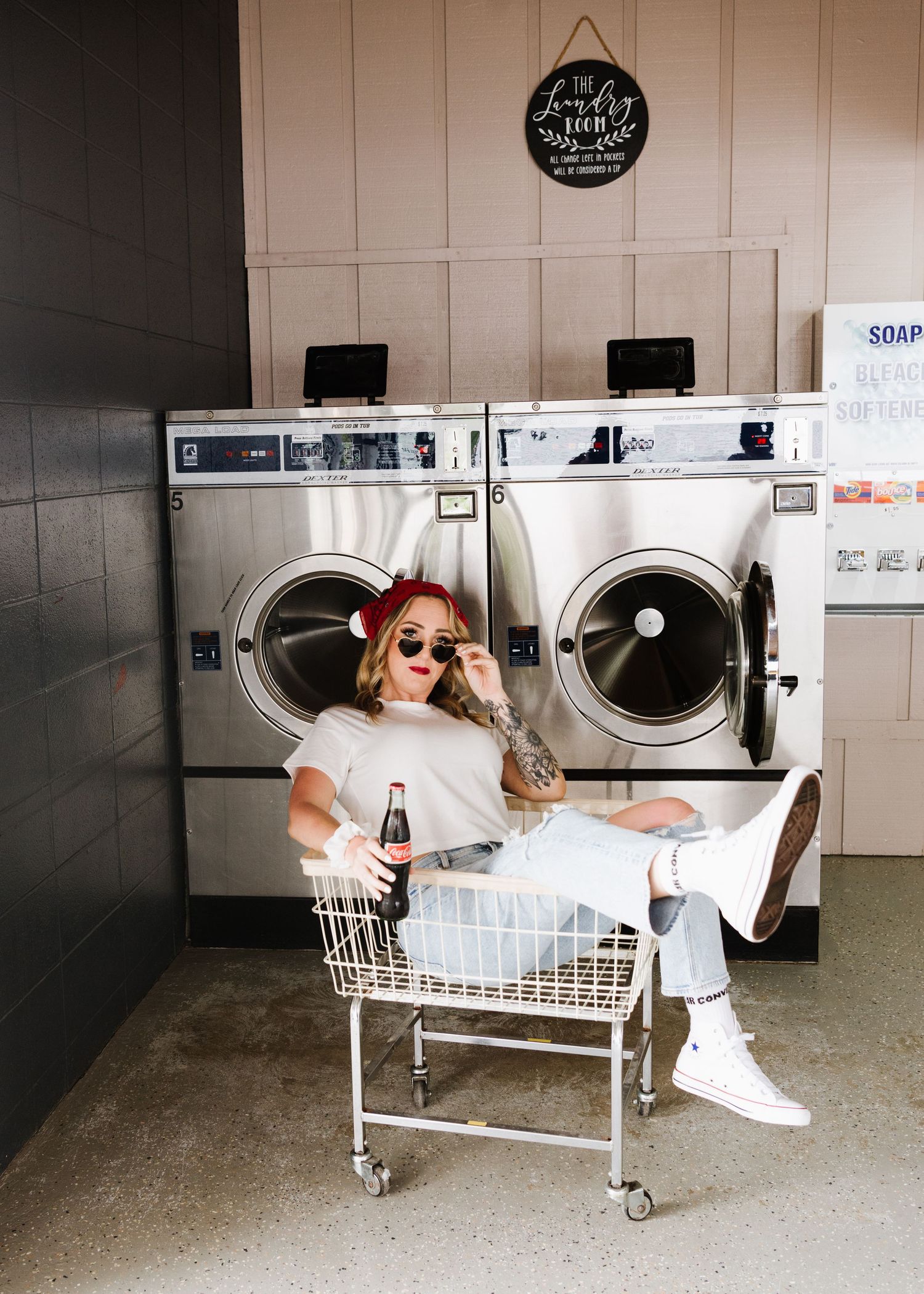 Someone sits on a laundry cart in a retro-style laundromat with vintage washing machines and a rustic wall sign.