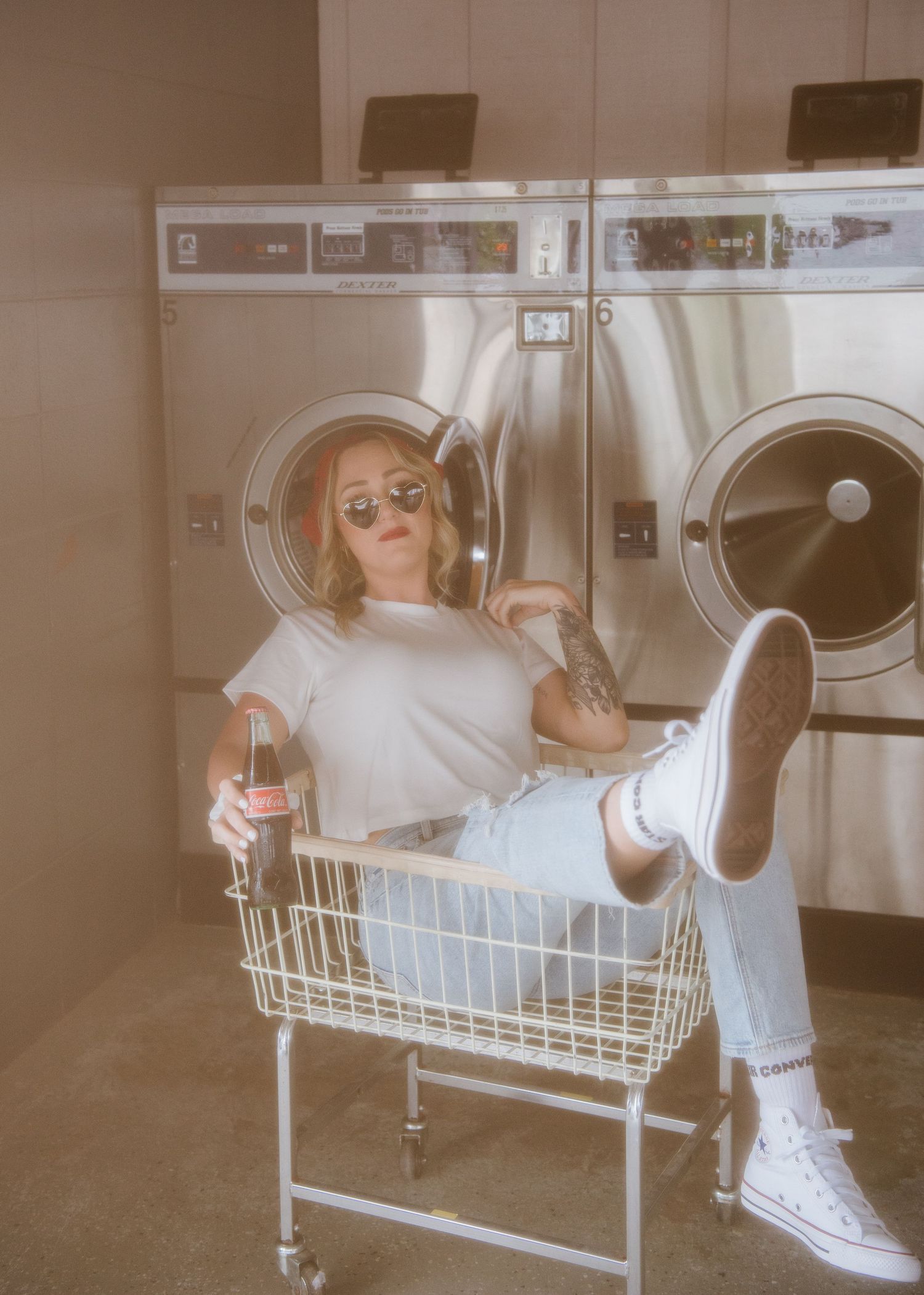 Retro-filtered laundromat scene showing figure lounging in shopping cart with wine glass against washing machines.
