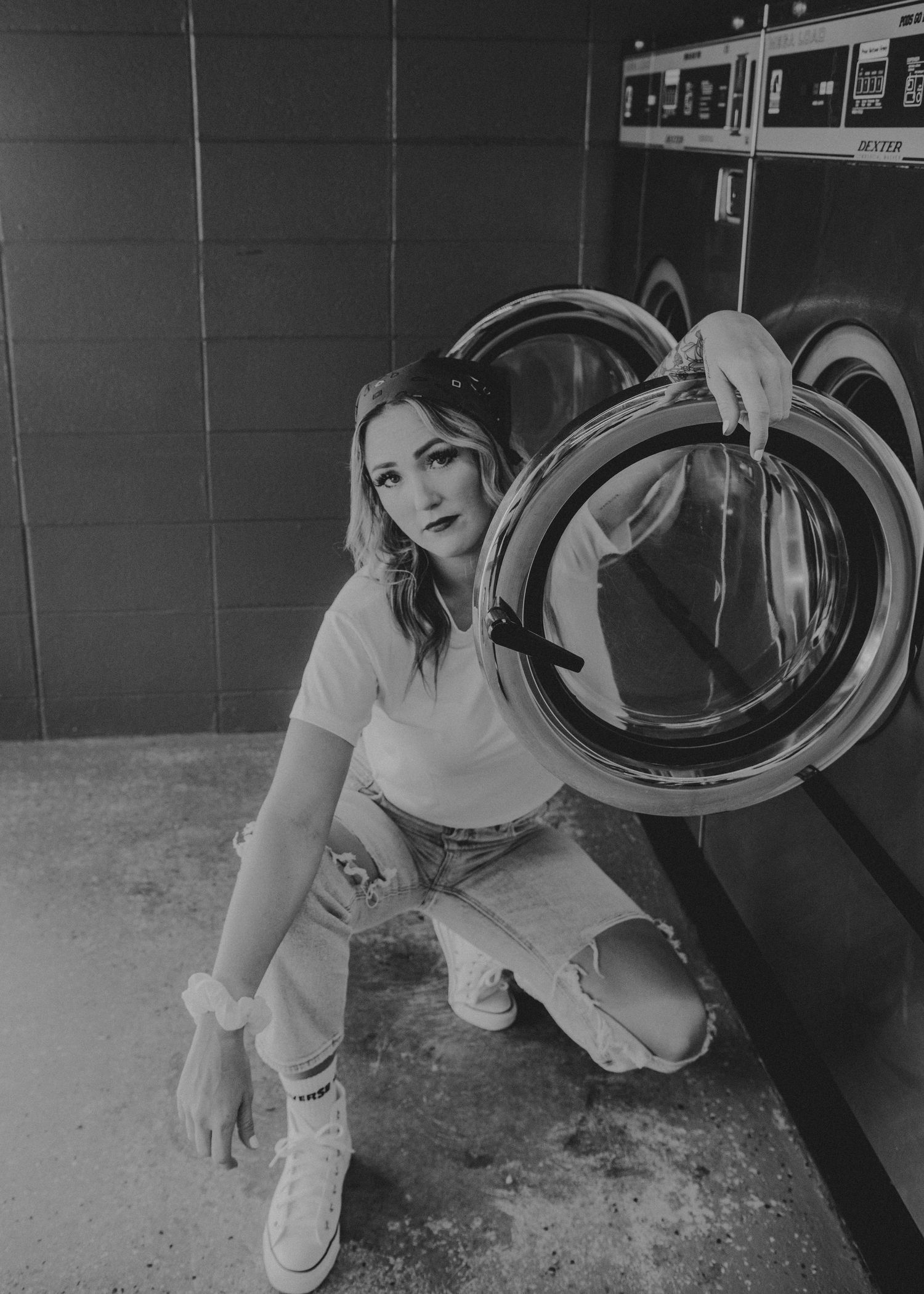 Black and white photo at a laundromat showing someone crouched next to a washing machine wearing casual attire.