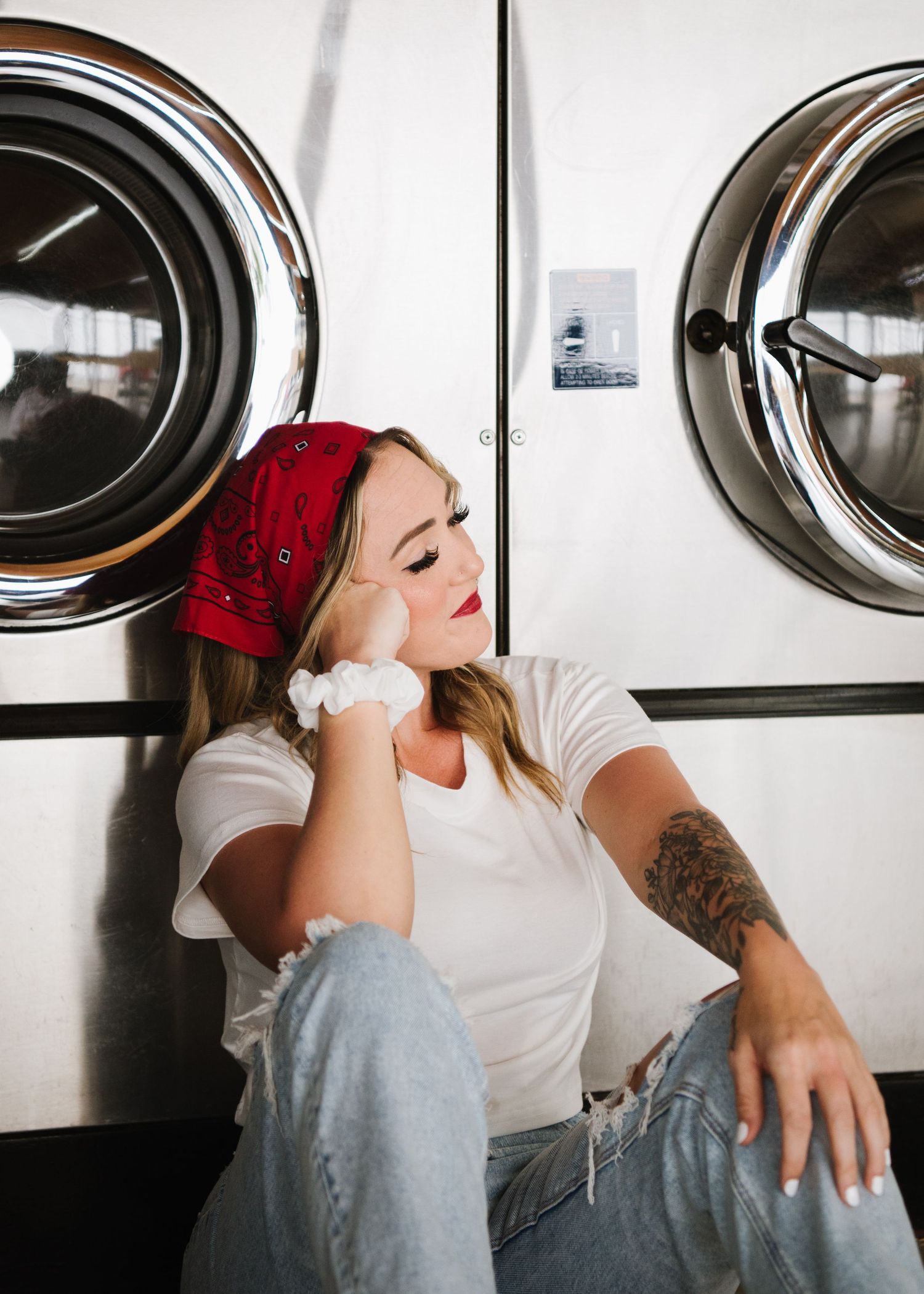 Person in red bandana and white t-shirt resting against industrial washing machines in a laundromat.
