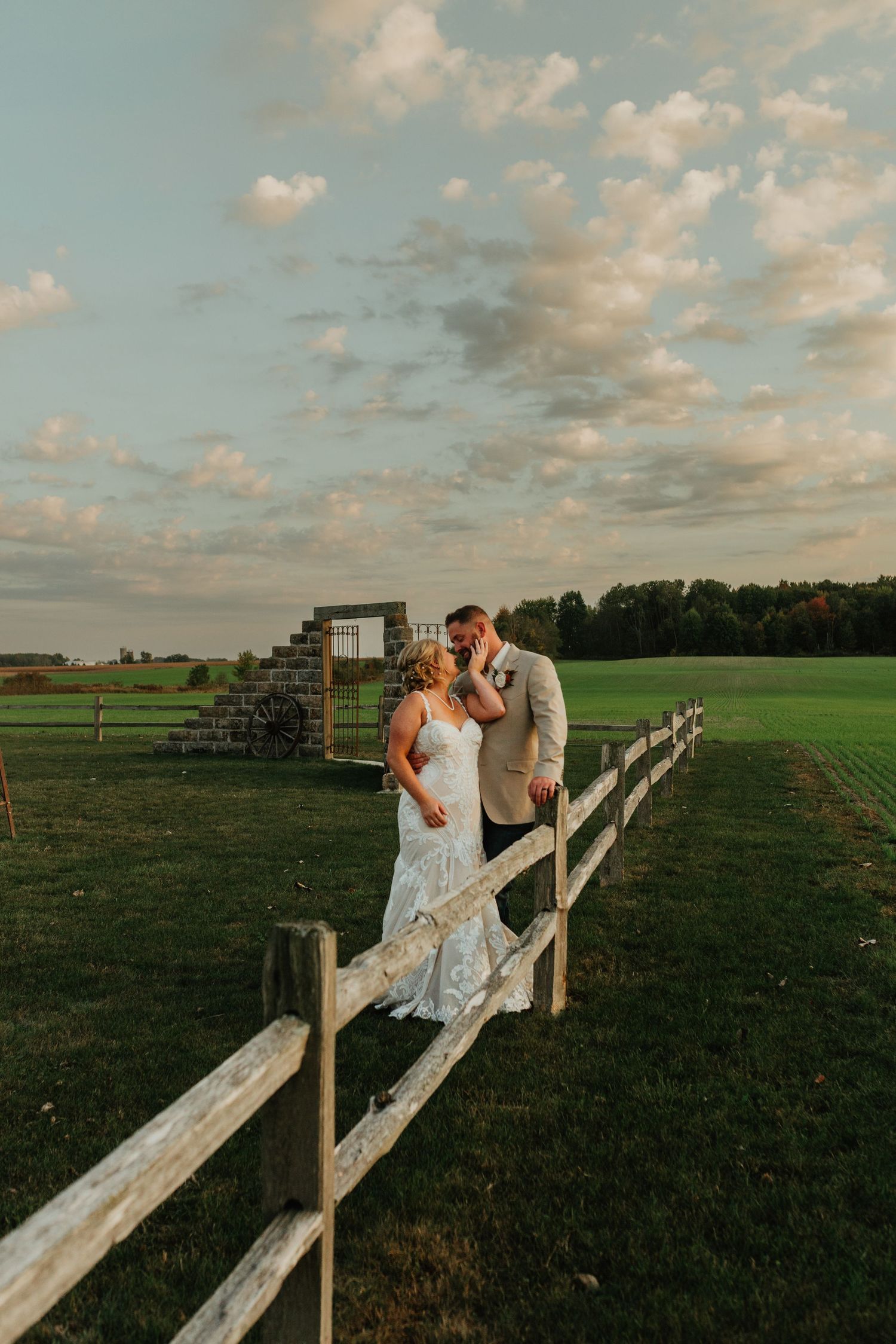Couple sharing a romantic moment on a rustic wooden fence at sunset with green fields and soft clouds in the background.